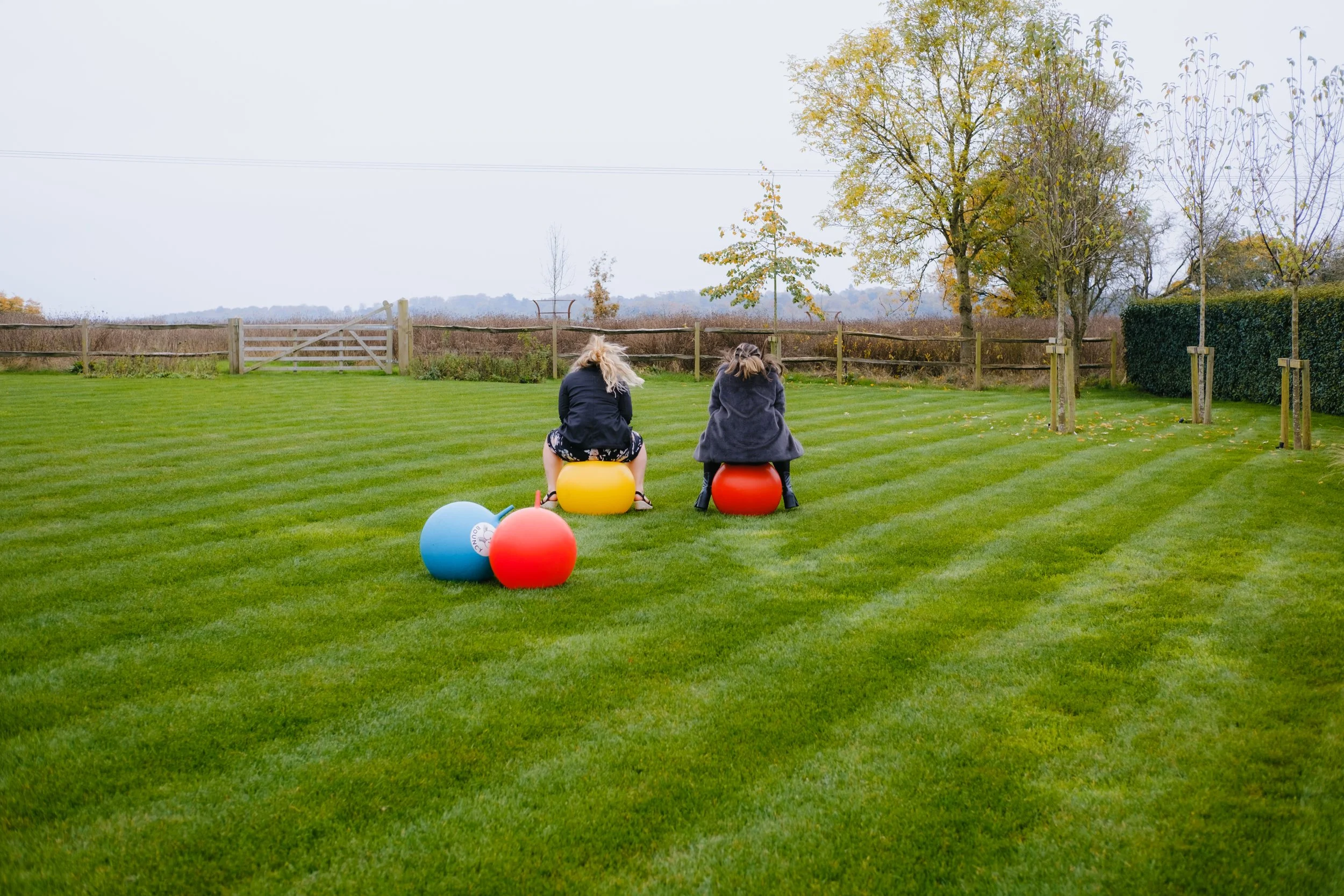 Two people sitting on yellow and red exercise balls on a grass lawn, with a blue exercise ball nearby, surrounded by trees and a wooden fence.