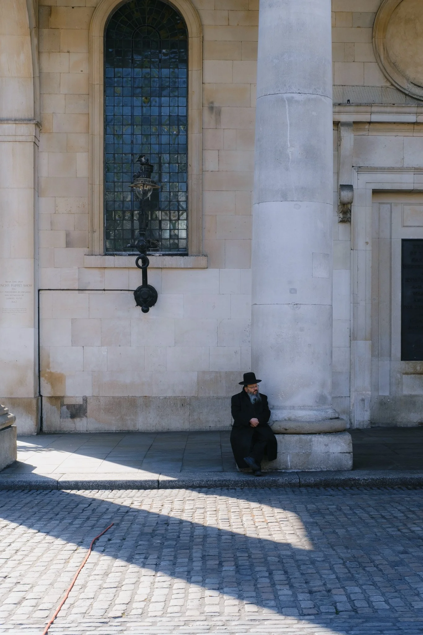 Man in black coat and hat sitting by stone pillar, with stone building and large window in background.