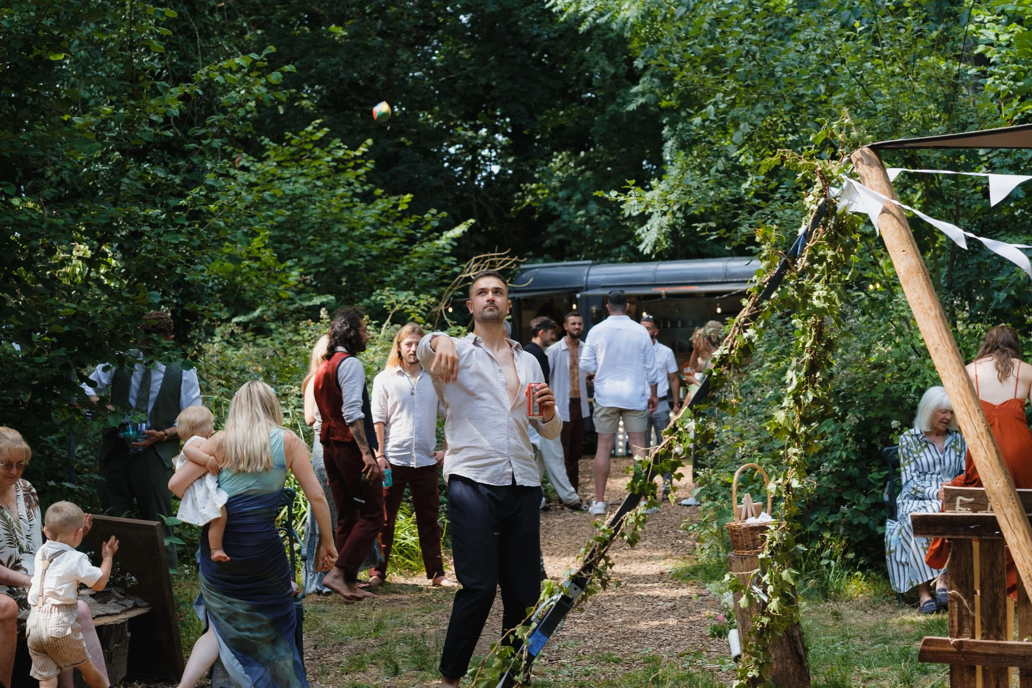 People gather in a lush outdoor wooded area for a celebration or event, with some standing and others sitting, and decorations like bunting strung on a wooden arch.