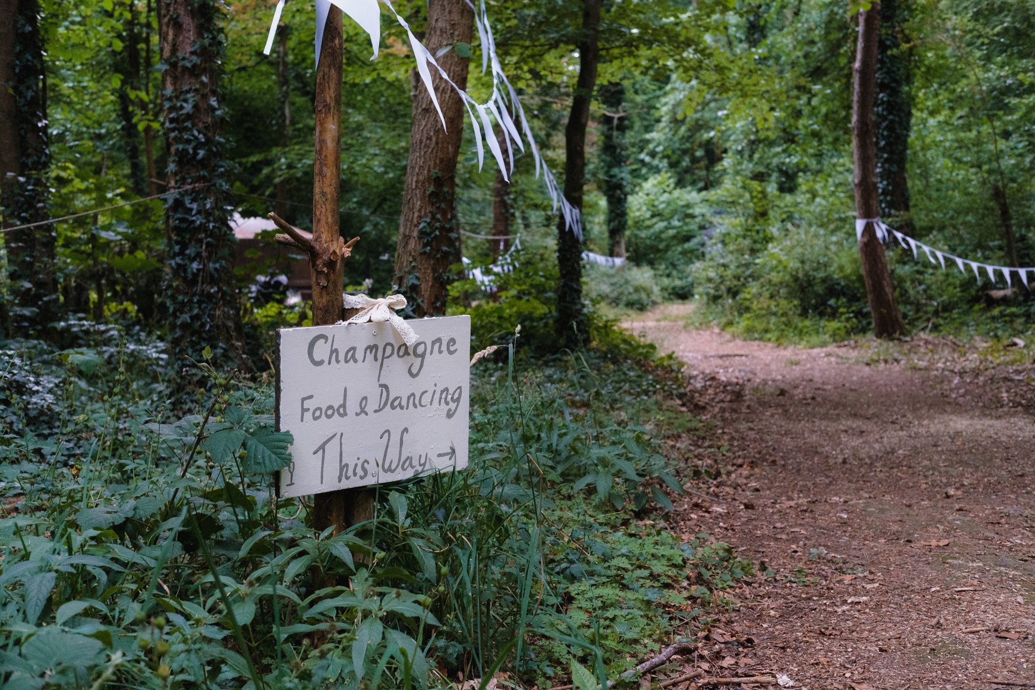 A wooded outdoor area with a dirt path and decorative white bunting hanging between trees. A sign on a tree reads 'Champagne Food & Dancing This Way' with an arrow pointing to the right.