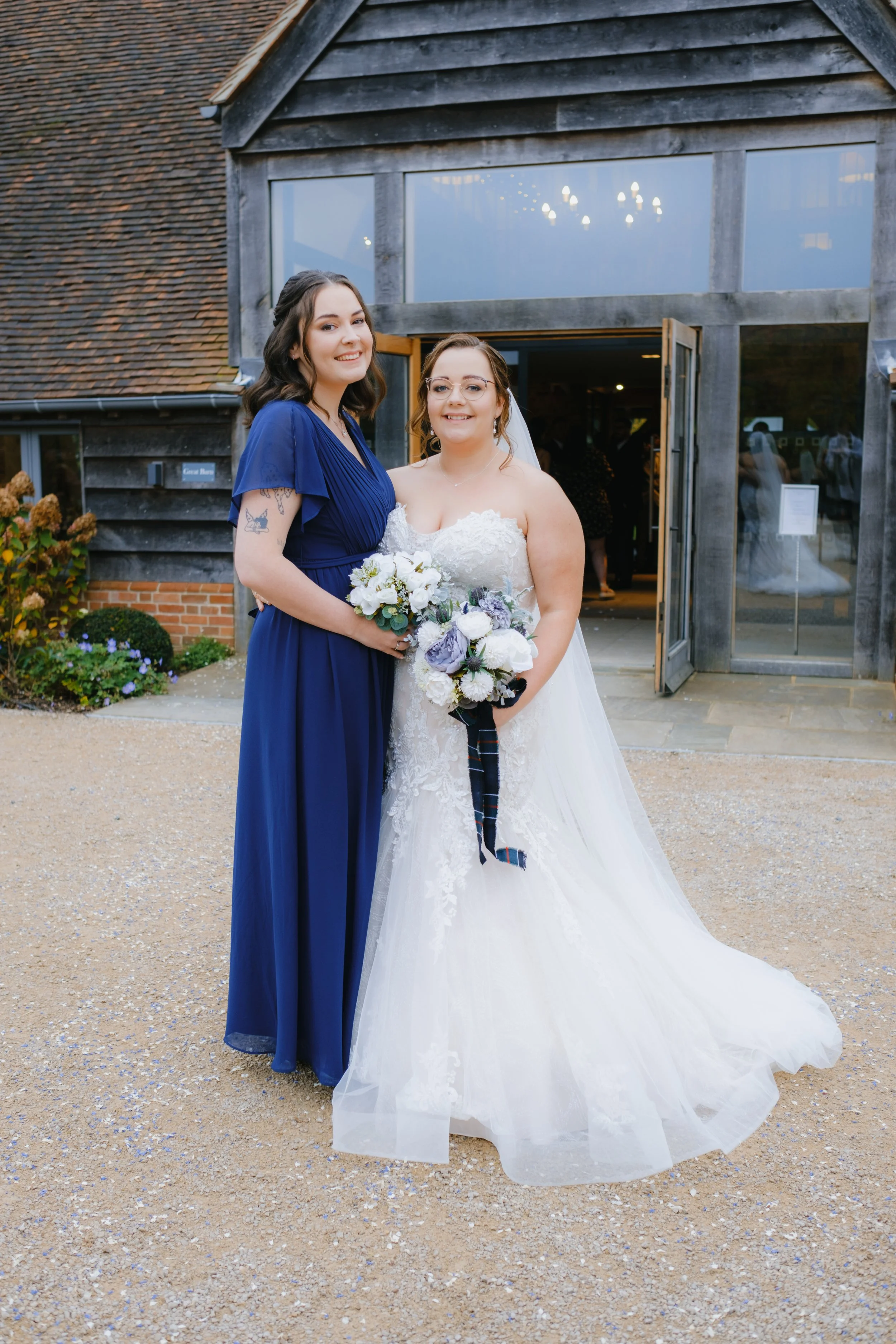 A bride in a white wedding dress and a bridesmaid in a blue dress standing in front of a rustic building with bouquets of flowers.