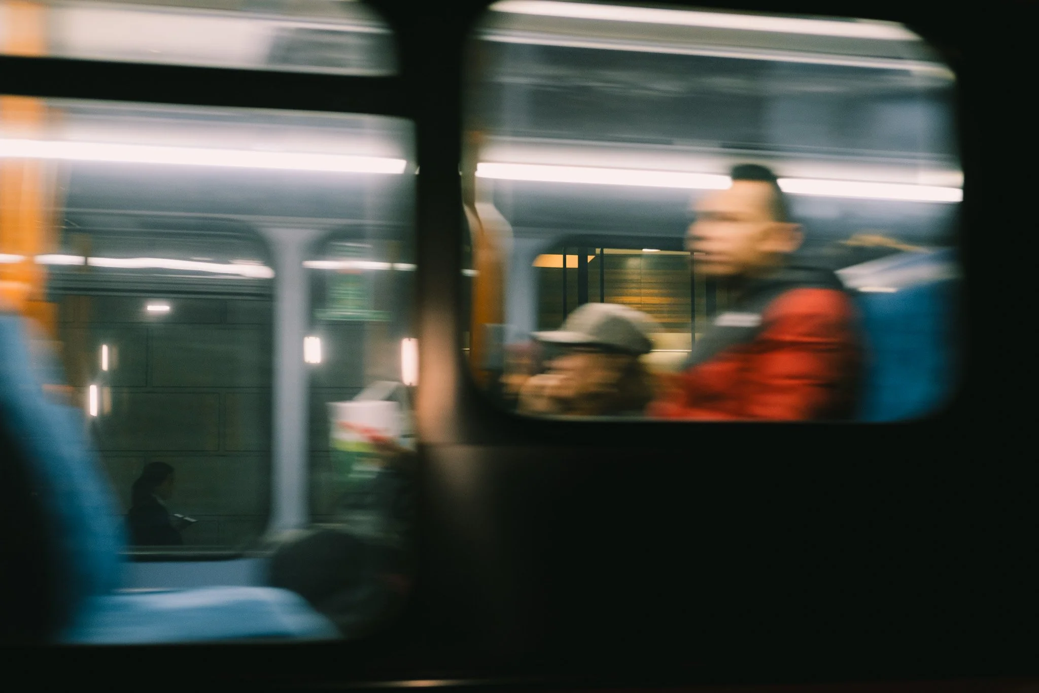 Blurry view from a train window showing people sitting and standing inside, with motion blur suggesting movement.
