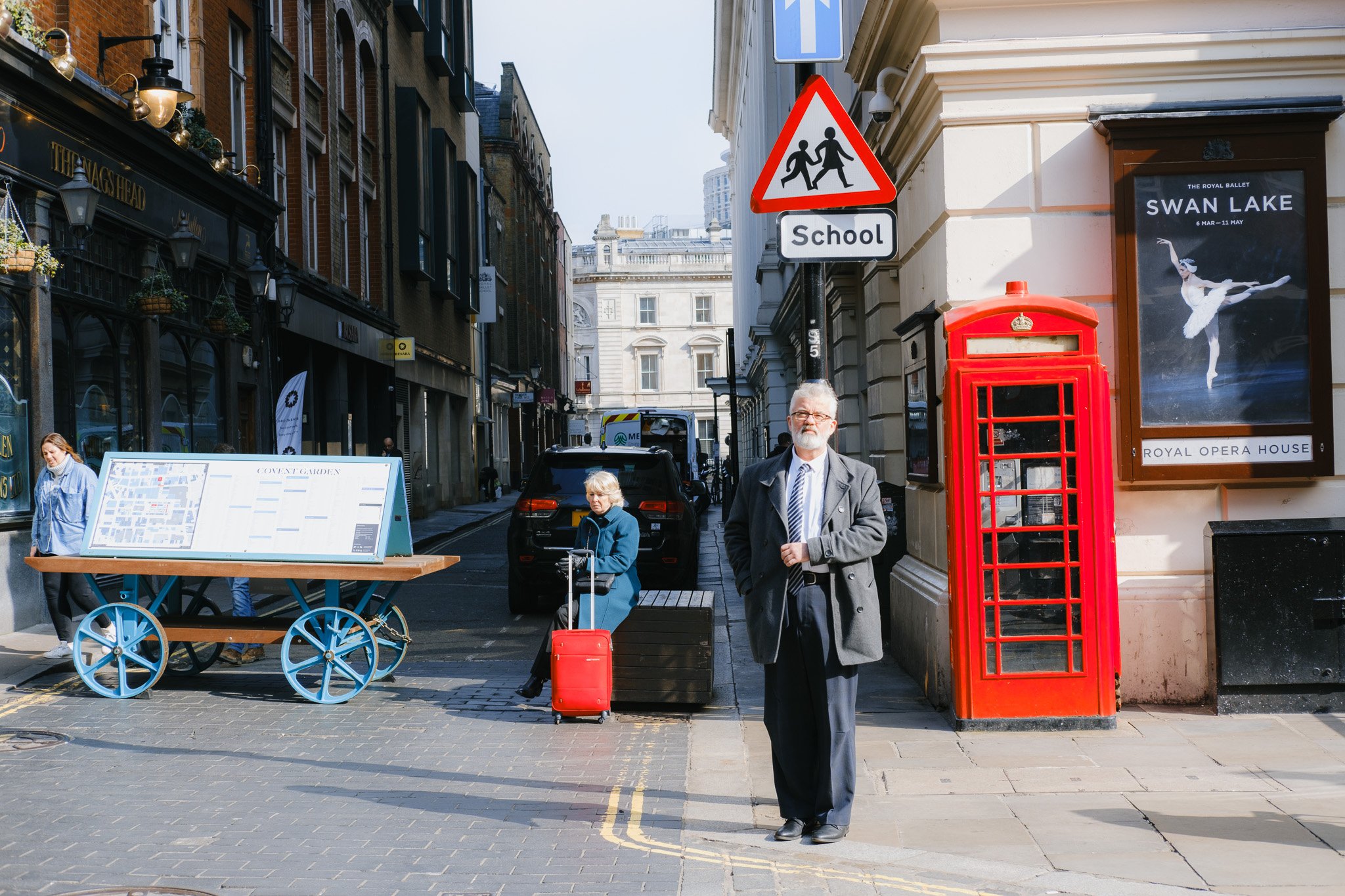 Street scene in Covent Garden, London with a red phone booth, 'School' road sign, Swan Lake poster, and people walking.
