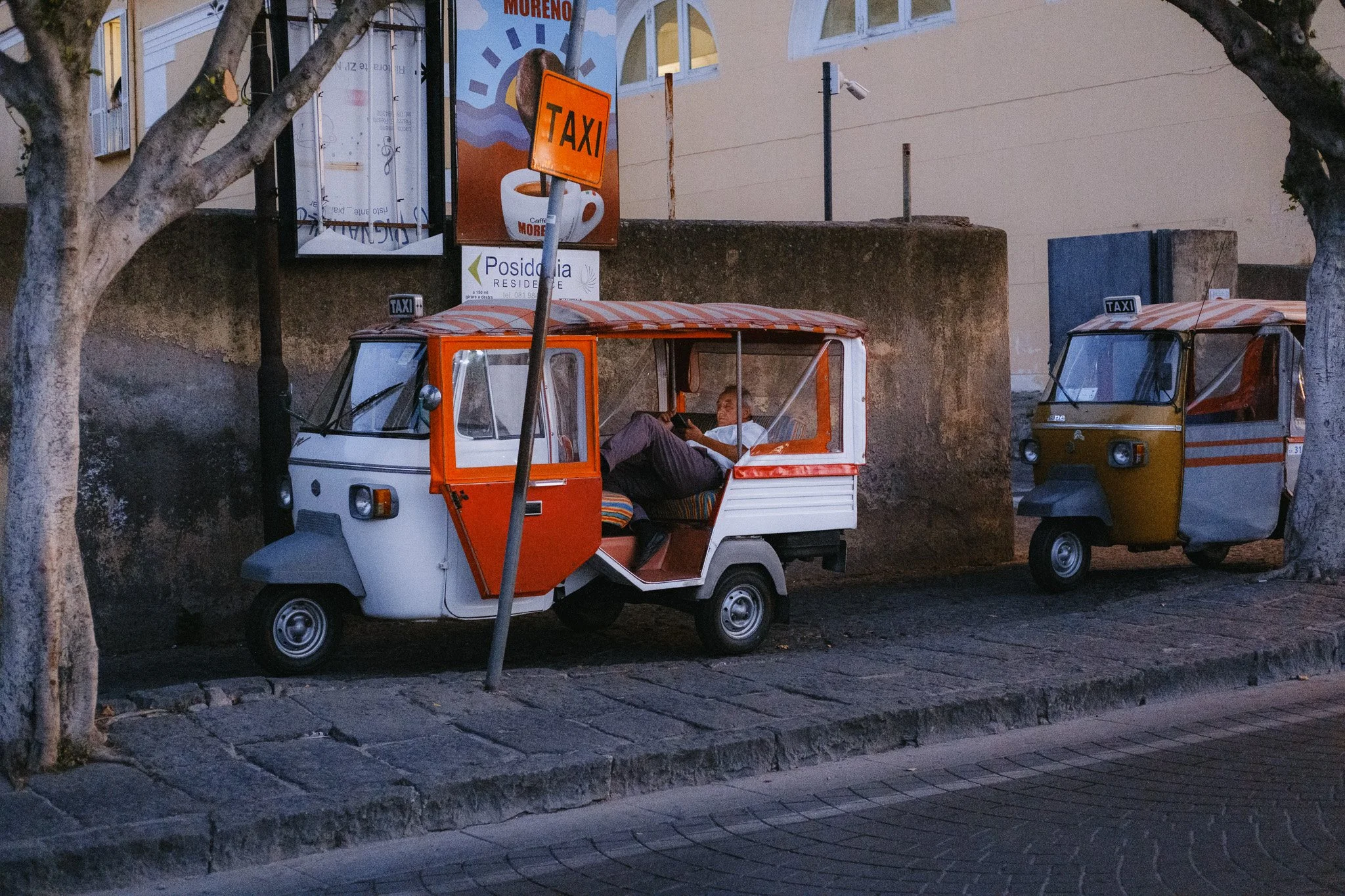 Two tuk-tuks parked under trees, one red and white with a person inside, and a yellow one behind, with a taxi sign above.