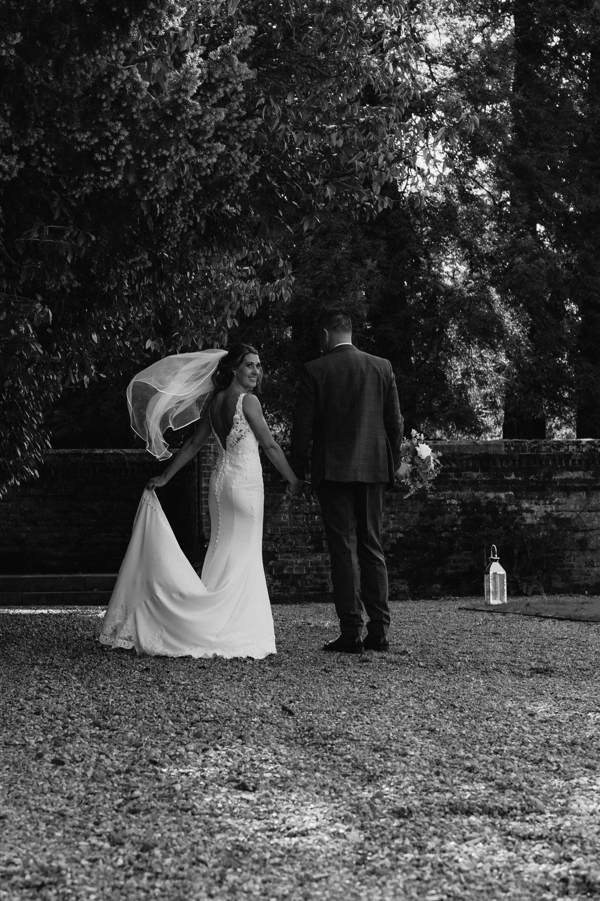 Black and white photo of a bride and groom holding hands, bride's veil flowing, outdoors with trees in background.