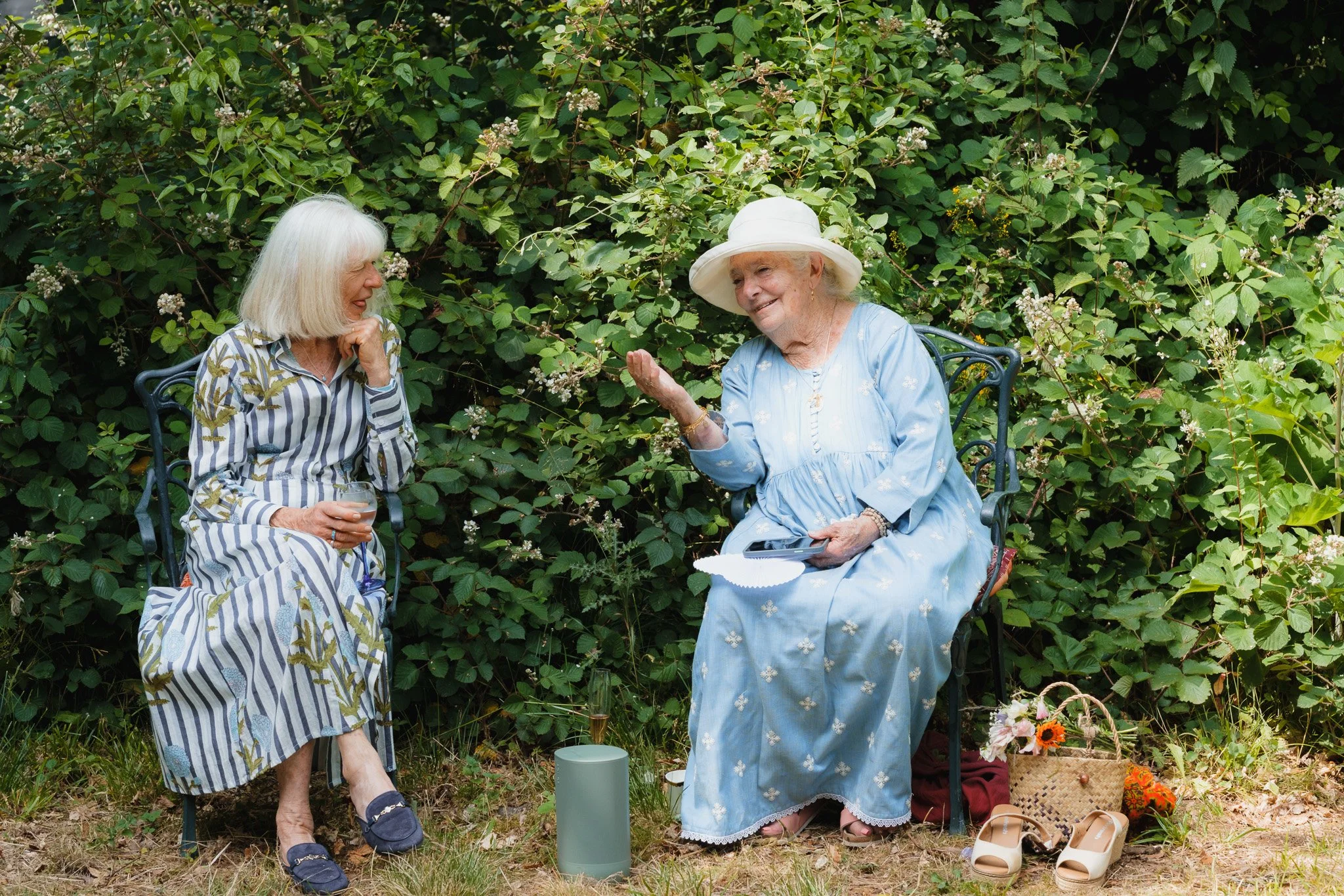 Two elderly women sit outdoors in front of a bush, enjoying a conversation. One woman wears a striped dress and holds a glass, while the other wears a light blue dress and a wide-brimmed hat, gesturing with her hand. There are flowers, a basket, shoe
