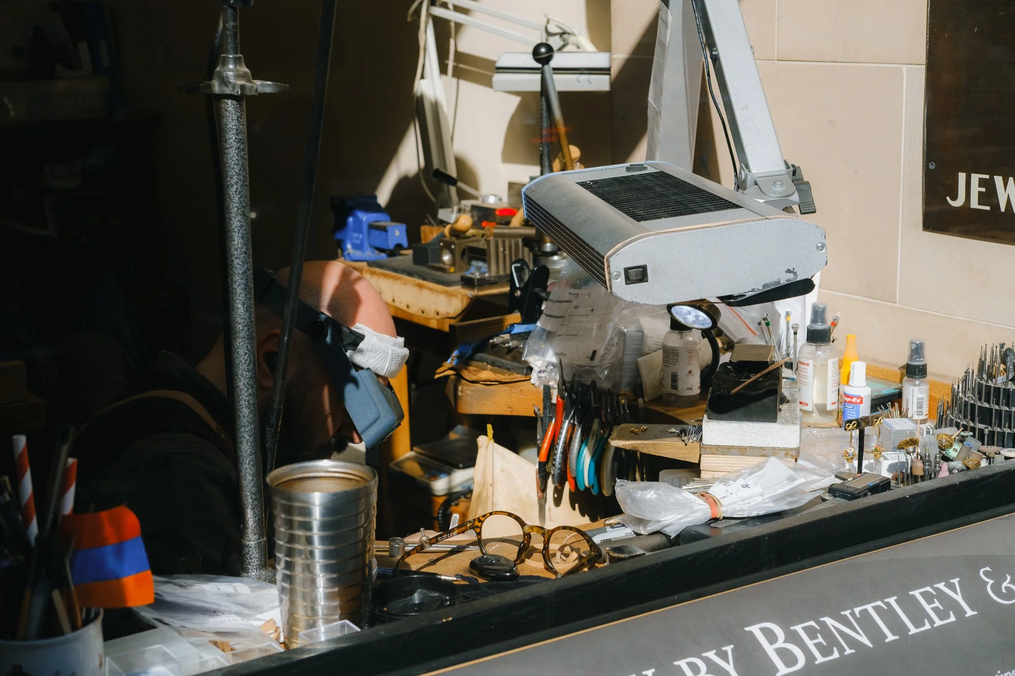 Jeweler working at a cluttered workbench with tools, magnifying glasses, and various equipment in a workshop.