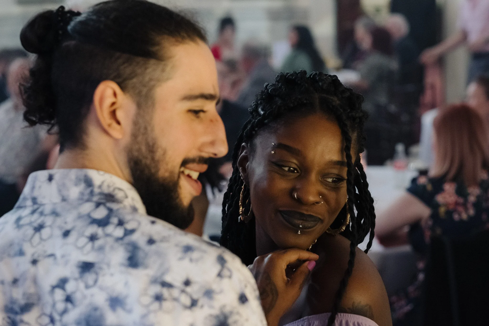 A smiling couple enjoying a social gathering, with the man having a beard and styled hair, and the woman having braided hair and hoop earrings.