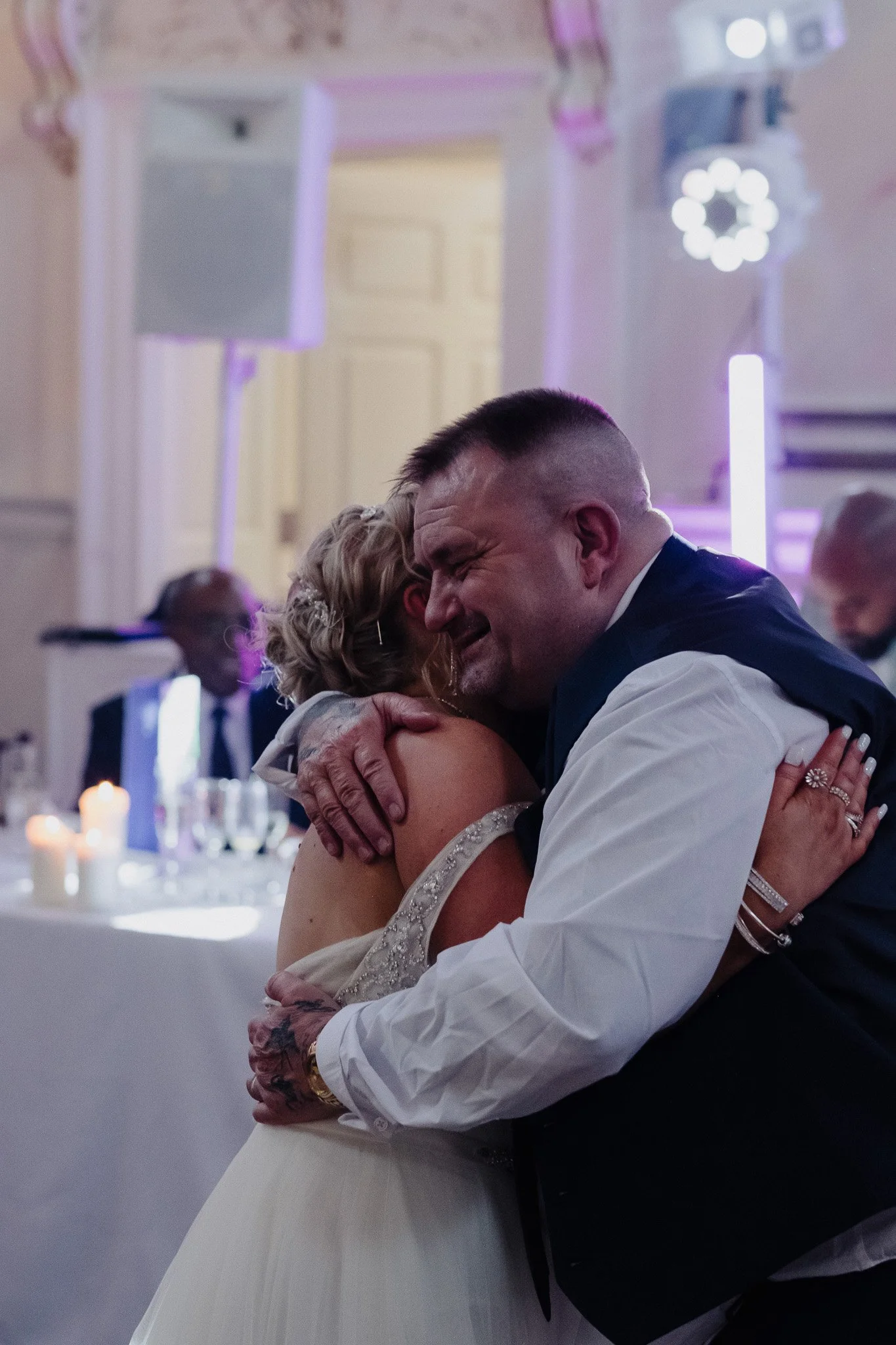 A man and woman hugging at a formal event, with candles and a decorated table in the background.