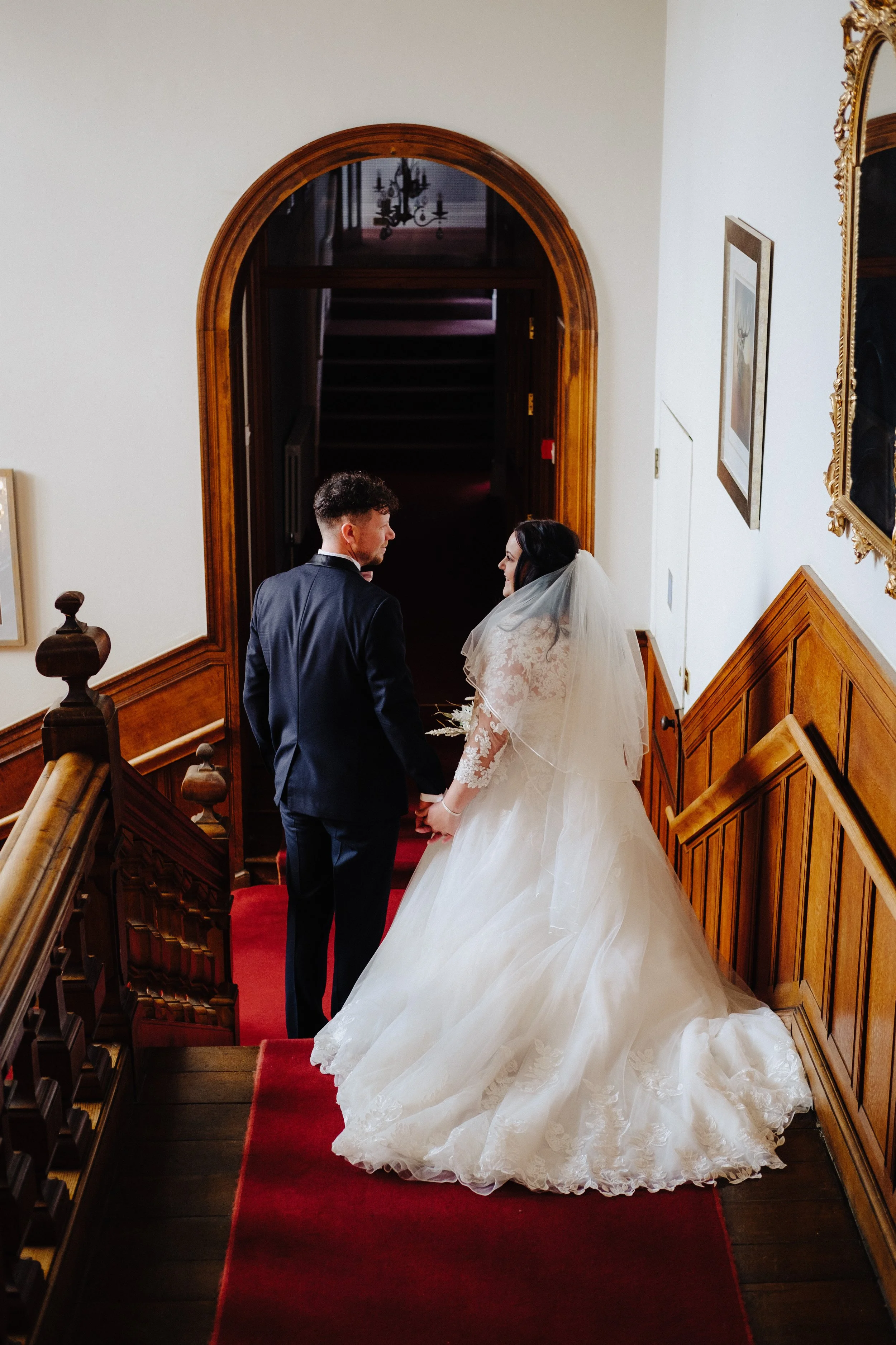 Bride and groom holding hands on a staircase, bride in a white gown, groom in a black suit, in a wood-paneled hallway with red carpet.