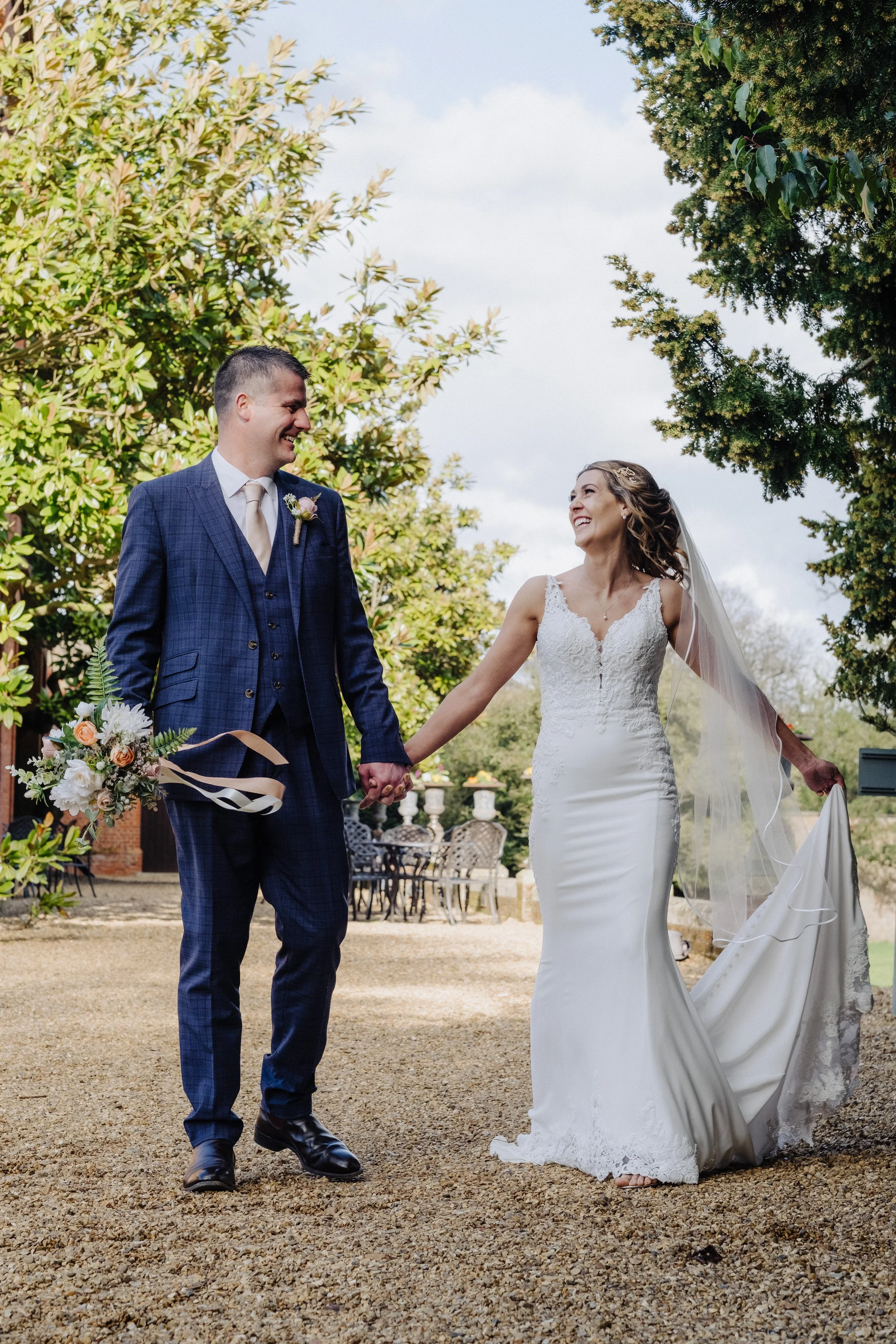 Bride and groom smiling on a gravel pathway, holding hands. The groom is in a blue suit with a floral boutonniere, and the bride is in a white wedding dress with lace details, holding a bouquet and veil. Surrounded by greenery and trees.