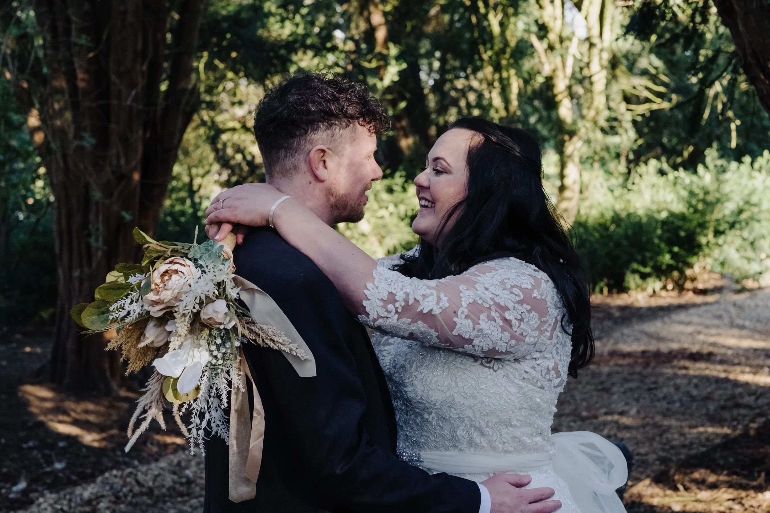 Bride and groom embracing in a forested setting, the bride holding a floral bouquet.