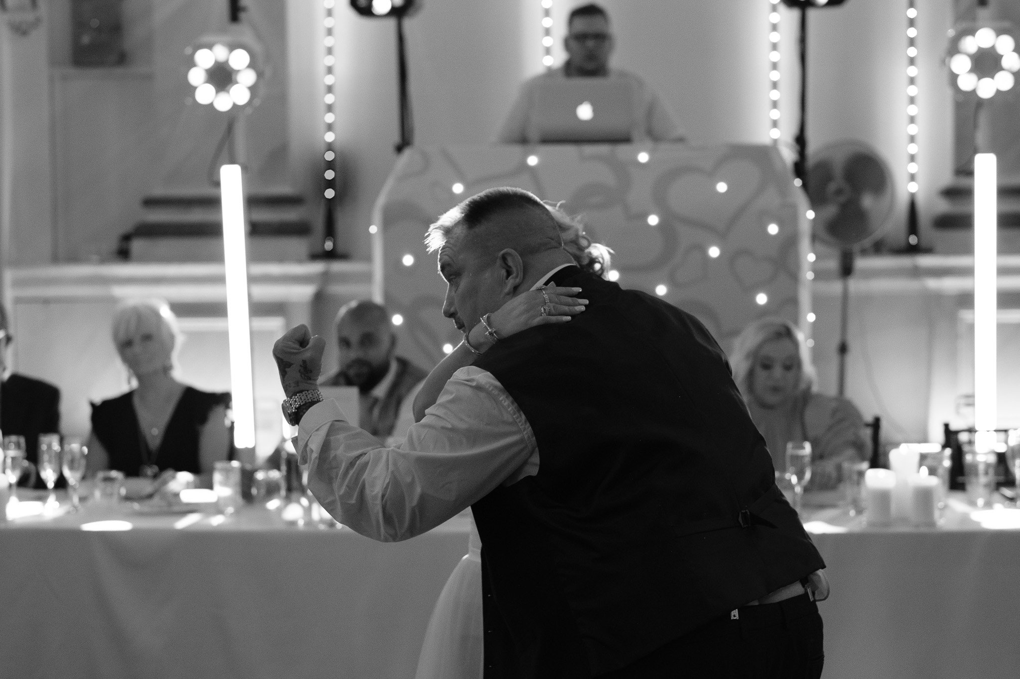Black and white photo of a man dancing at a wedding reception with guests seated at a table behind them.