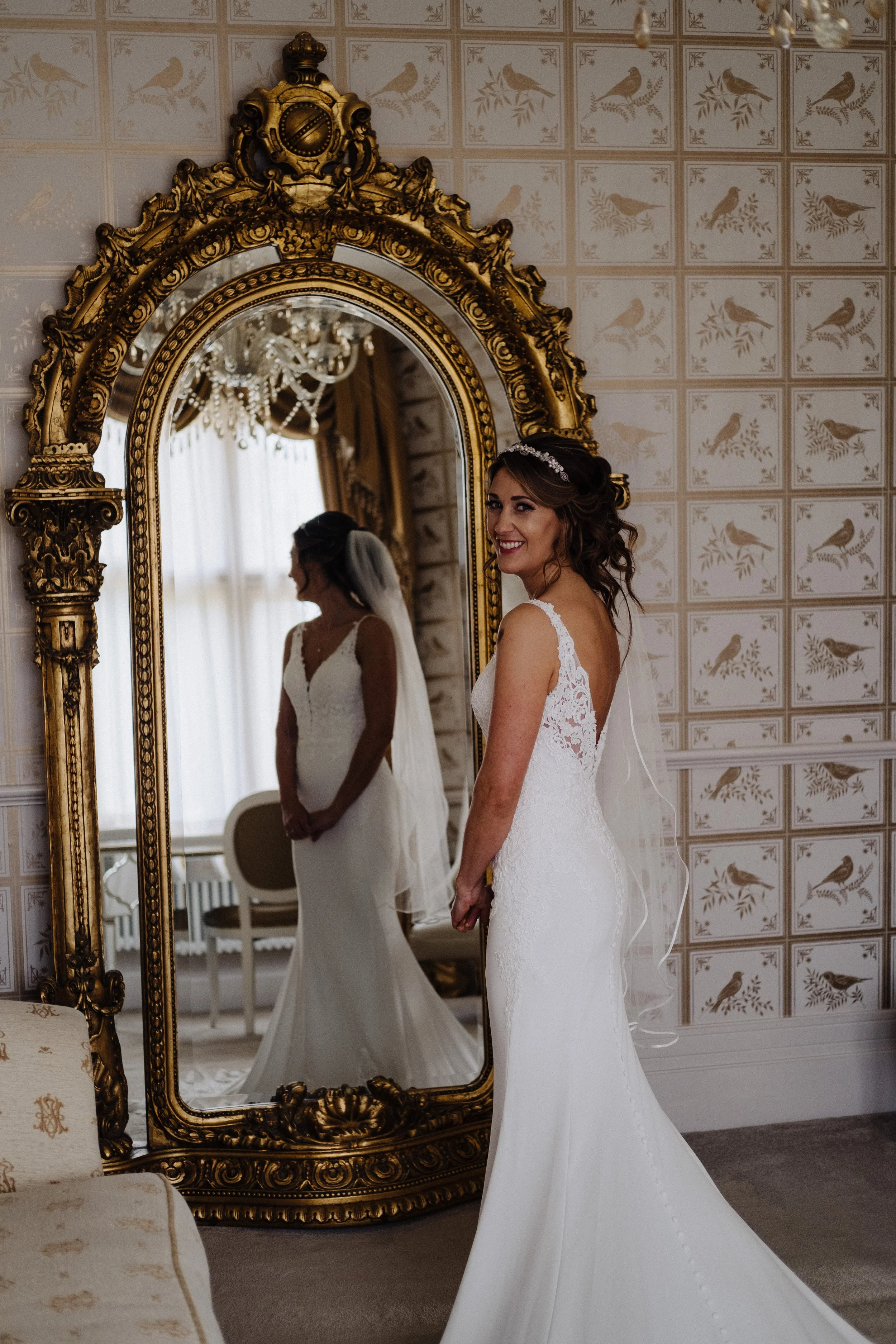 Bride in a white lace wedding dress standing in front of an ornate gold mirror, smiling with her reflection visible. The room has classic wallpaper with bird patterns, and a chandelier is partially visible.