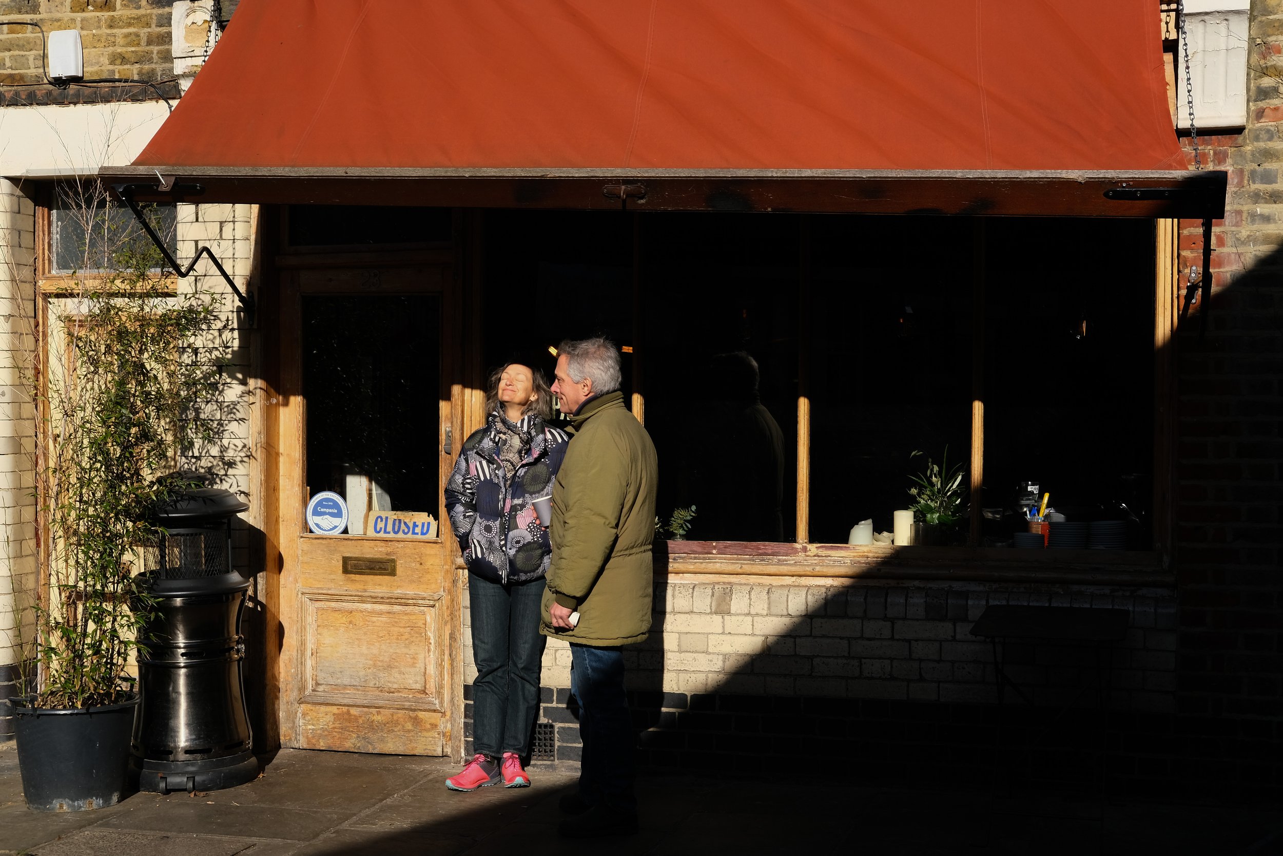 Two people standing in front of a closed shop with a red awning, one person looking upward with eyes closed.