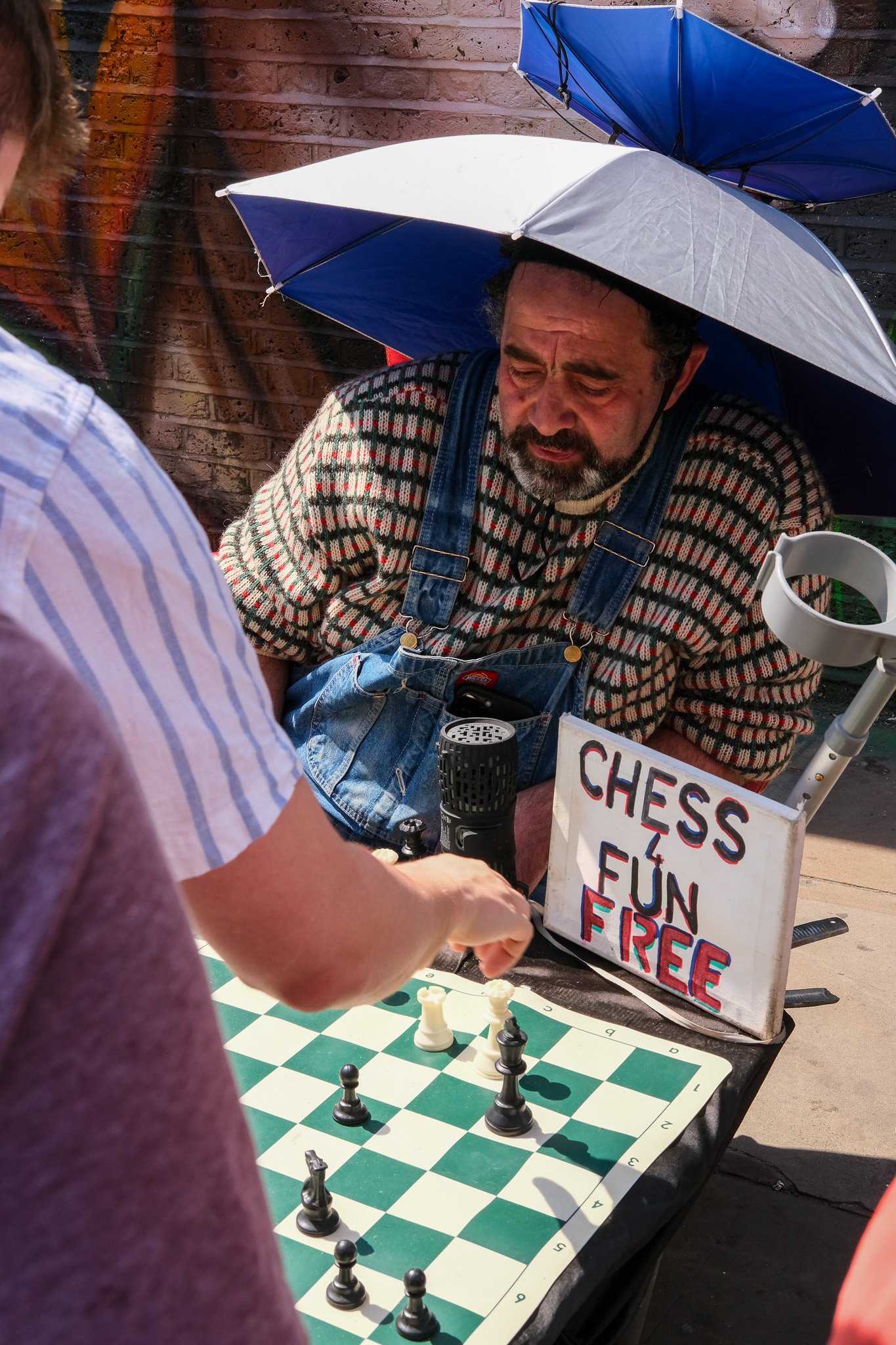 A man sitting under an umbrella playing chess on a sidewalk with a sign that says, "Chess 4 Fun Free." A person in front of him is moving a chess piece on the board.
