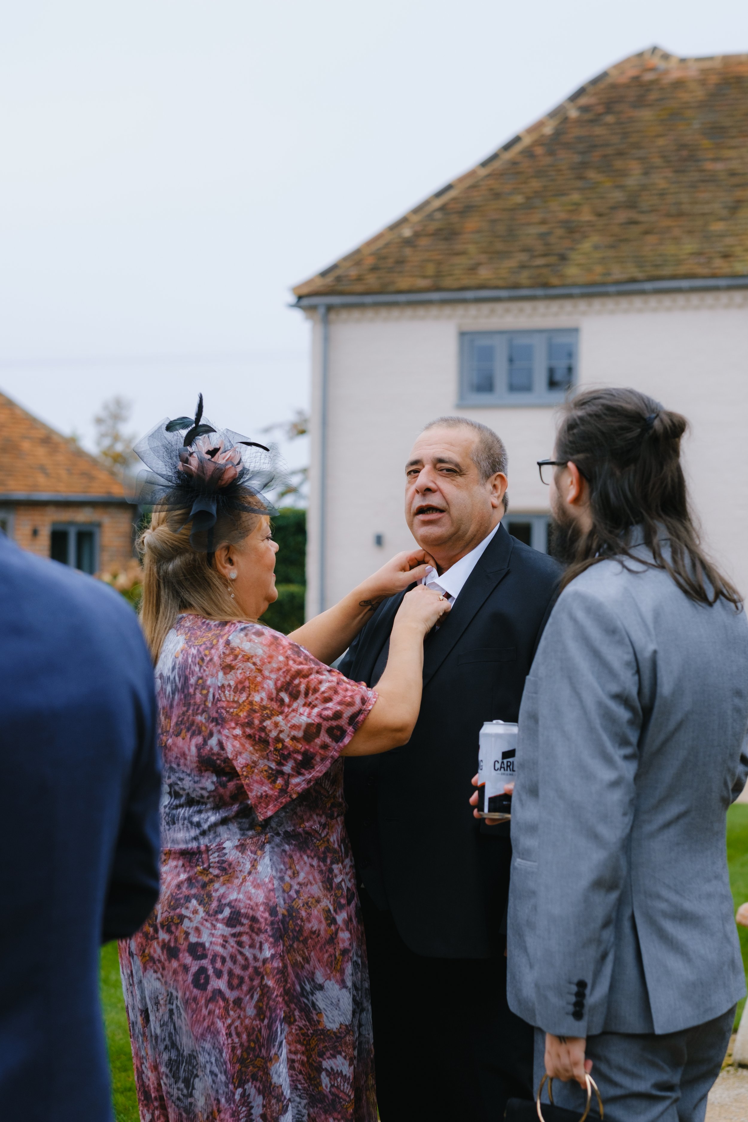 A woman adjusts a man's tie outdoors, surrounded by people dressed in formal attire, with a house in the background.