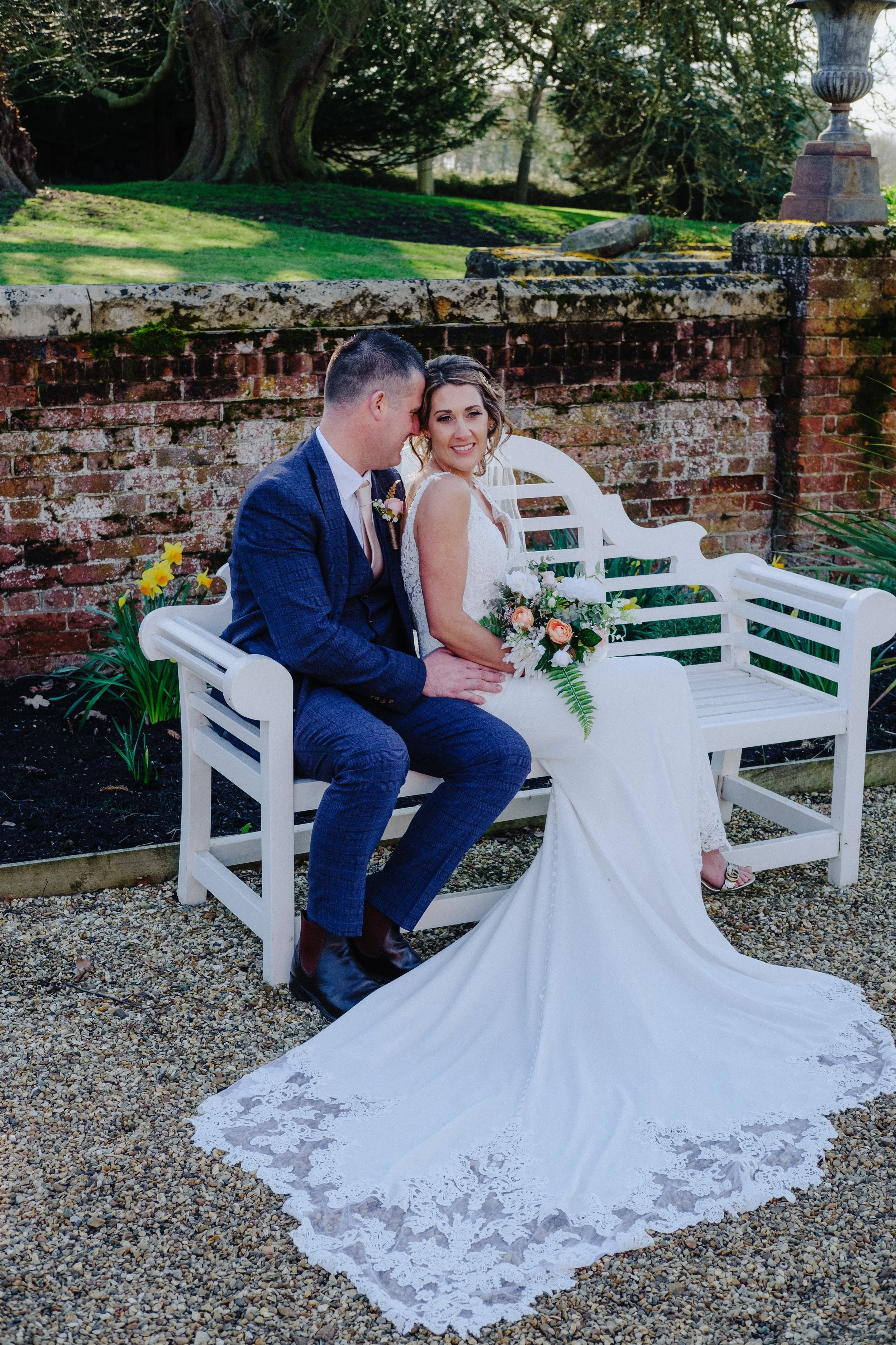 Bride and groom sitting on a white bench in a garden, with the bride holding a bouquet and wearing a white dress, and the groom in a navy blue suit.