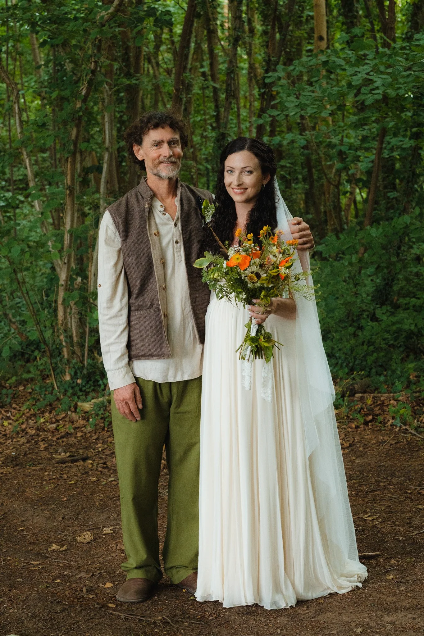 A man and a woman in wedding attire standing in a wooded outdoor setting, smiling and holding a bouquet of flowers.