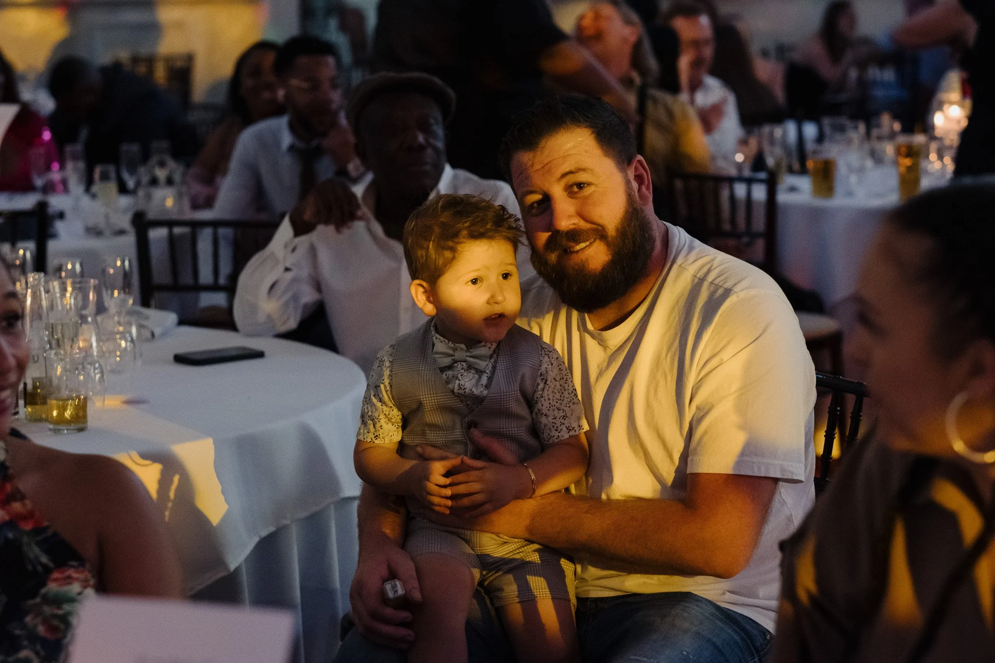 A man holding a child on his lap at a formal event, surrounded by people seated at tables with drinks.