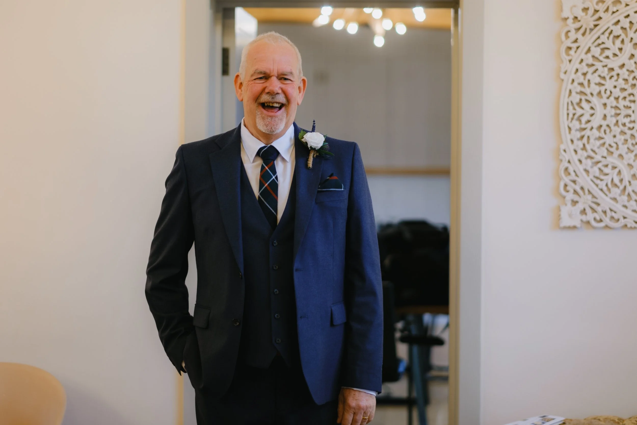 Smiling man in a navy suit with a white boutonnière standing indoors.
