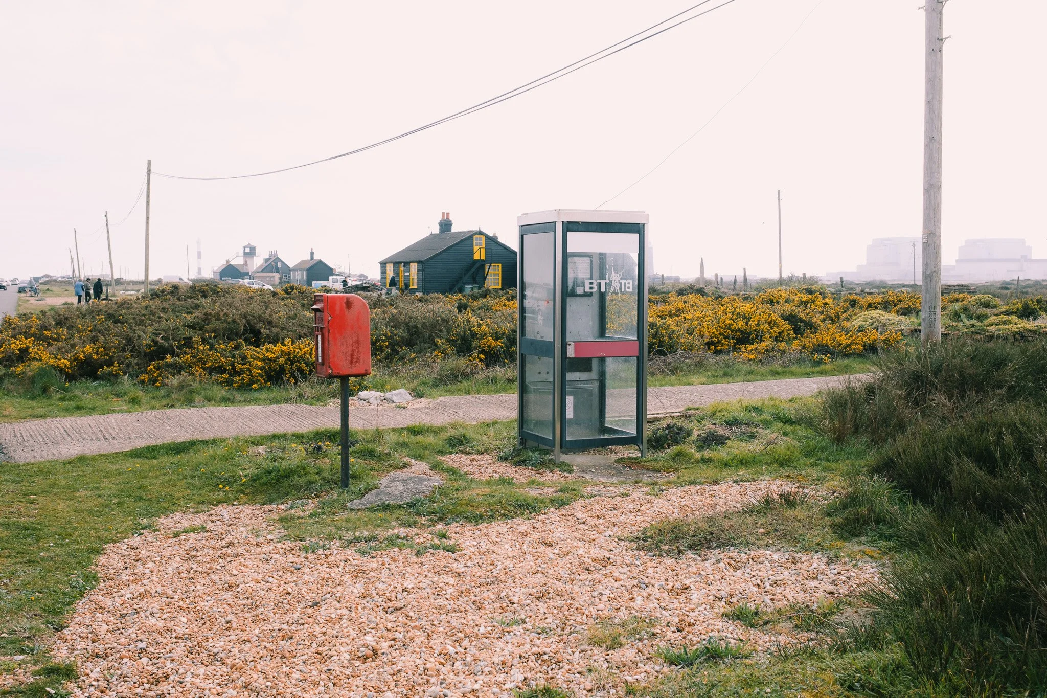 A rural landscape with a glass phone booth and red mailbox. The background features small houses, bushes with yellow flowers, and a cloudy sky. A gravel path and telephone poles are also visible in the scene.