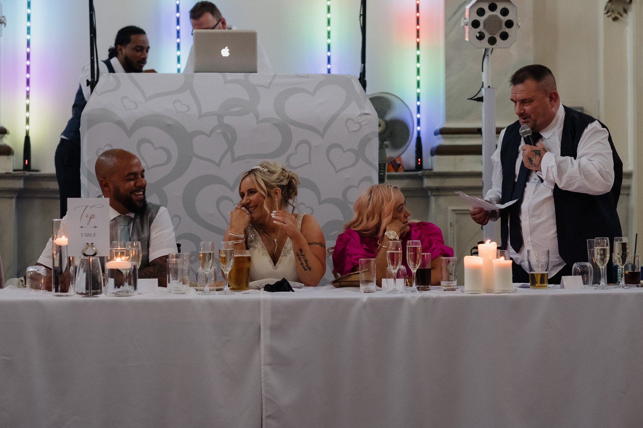 Wedding reception with a speaker and guests seated at a table with drinks and candles.