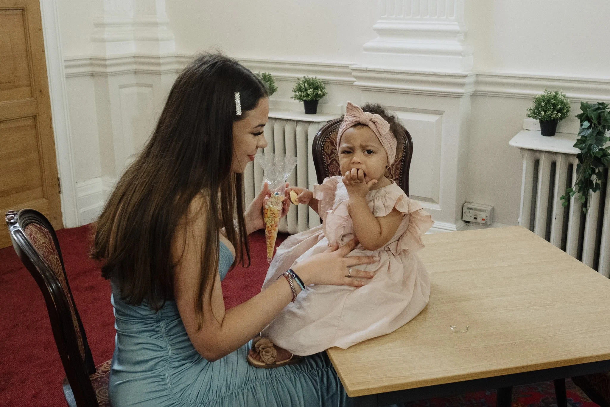 Woman in a blue dress interacting with a toddler in a pink dress seated on a table, with potted plants in the background.