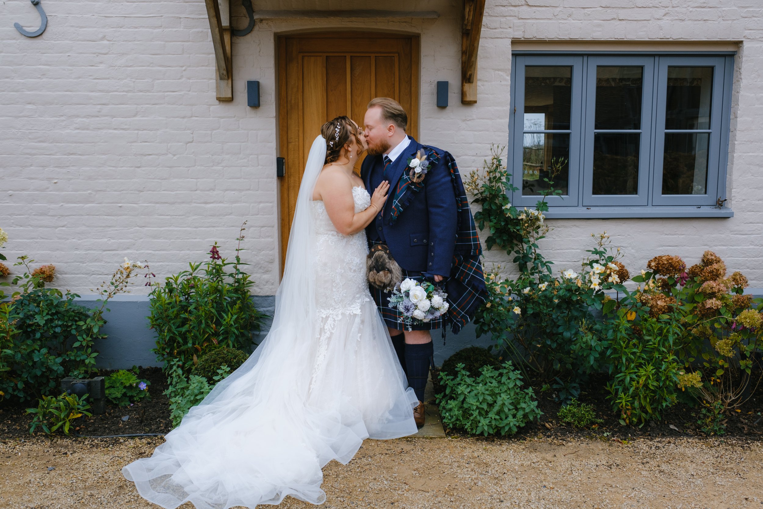 Bride in white dress and groom in traditional Scottish attire exchanging a kiss in front of a brick house with shrubs and flowers.