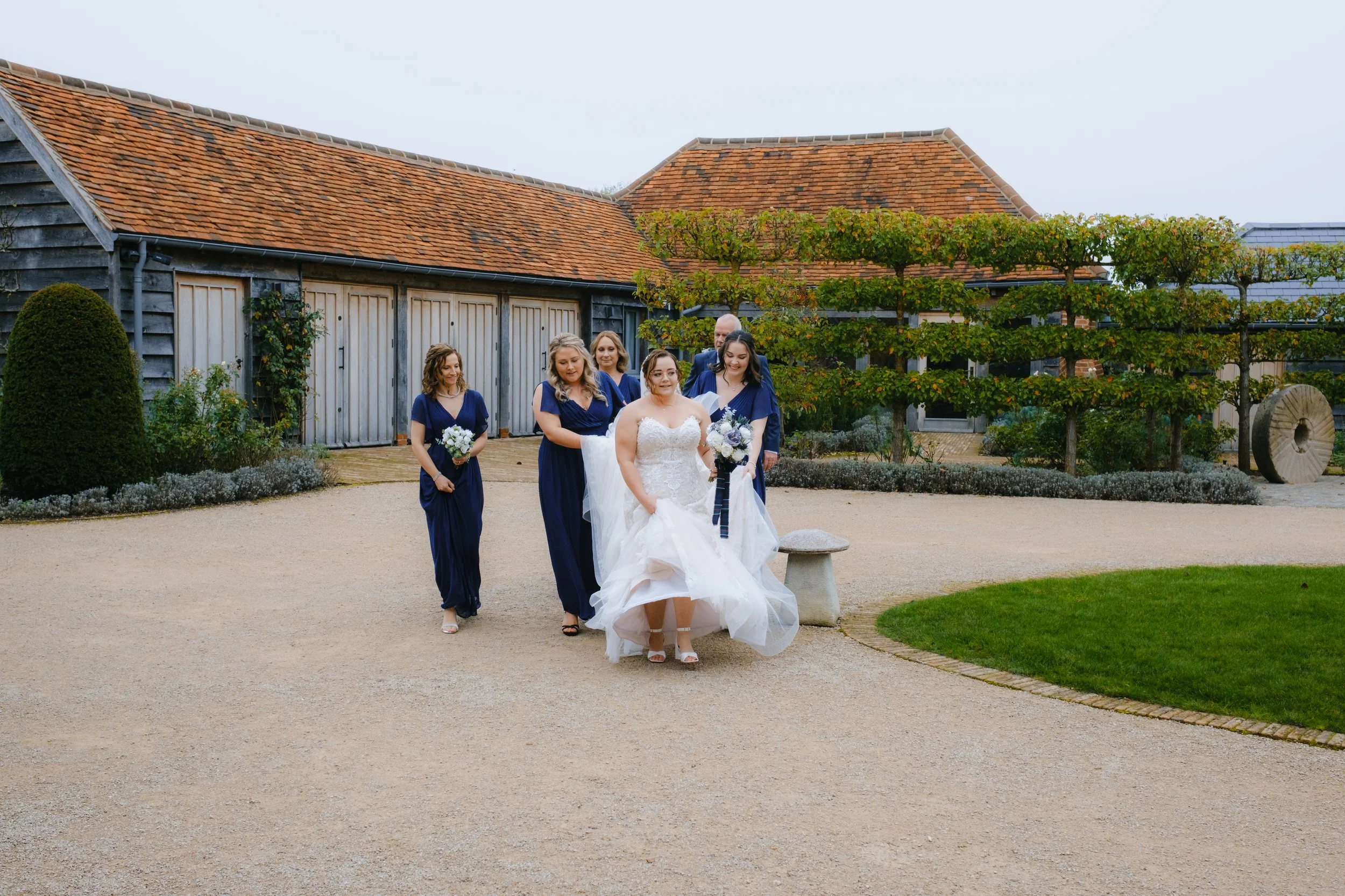 A bride in a white dress walks with bridesmaids in blue dresses and a man in a suit along a garden path in front of rustic buildings.