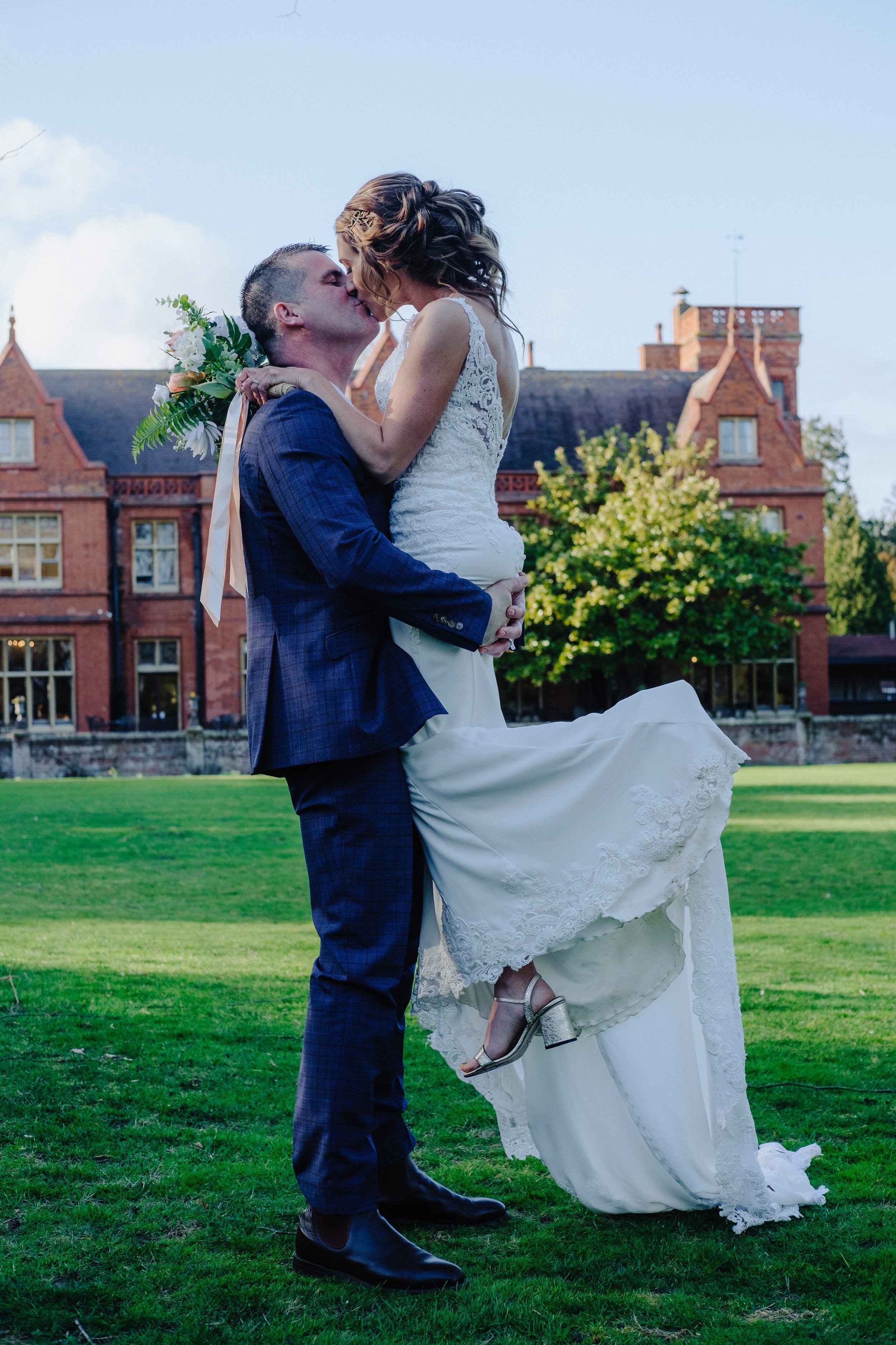 Wedding couple kissing outdoors, bride in white gown and groom in dark suit, historic building in background.