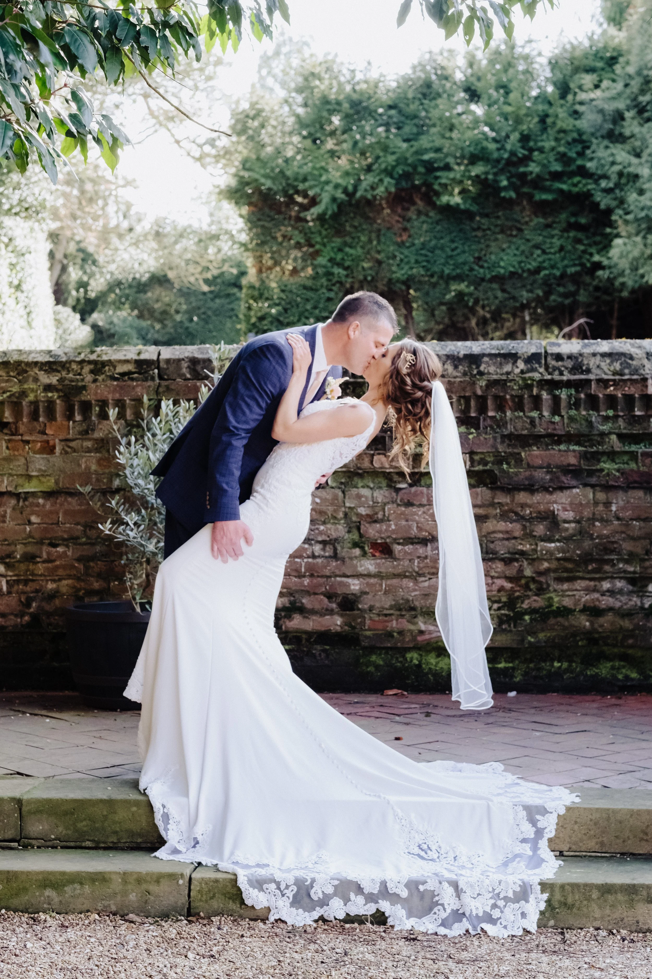 Bride and groom kissing in garden, bride in white dress with veil, groom in suit, stone steps in background.