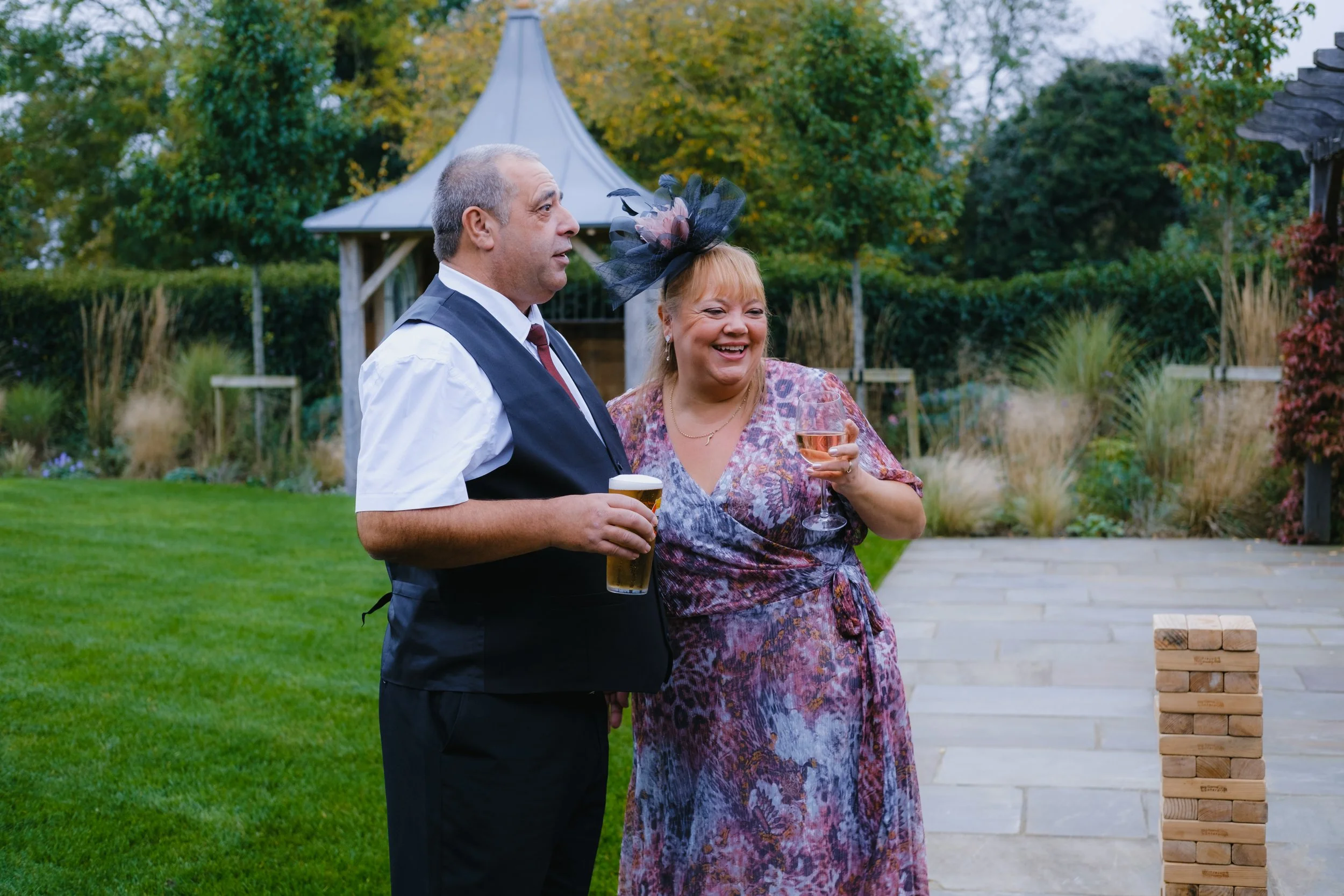 A man in a vest and tie holding a pint of beer stands next to a woman in a floral dress with a fascinator, holding a glass of wine, outdoors in a garden with a wooden gazebo in the background.