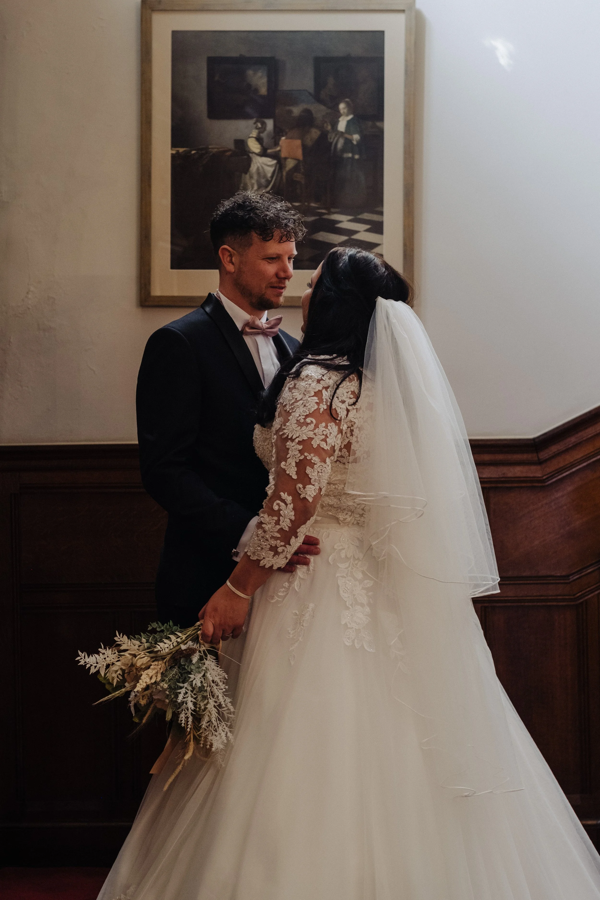 A bride and groom facing each other, with the bride holding a bouquet, standing in front of a framed painting on a wooden wall.