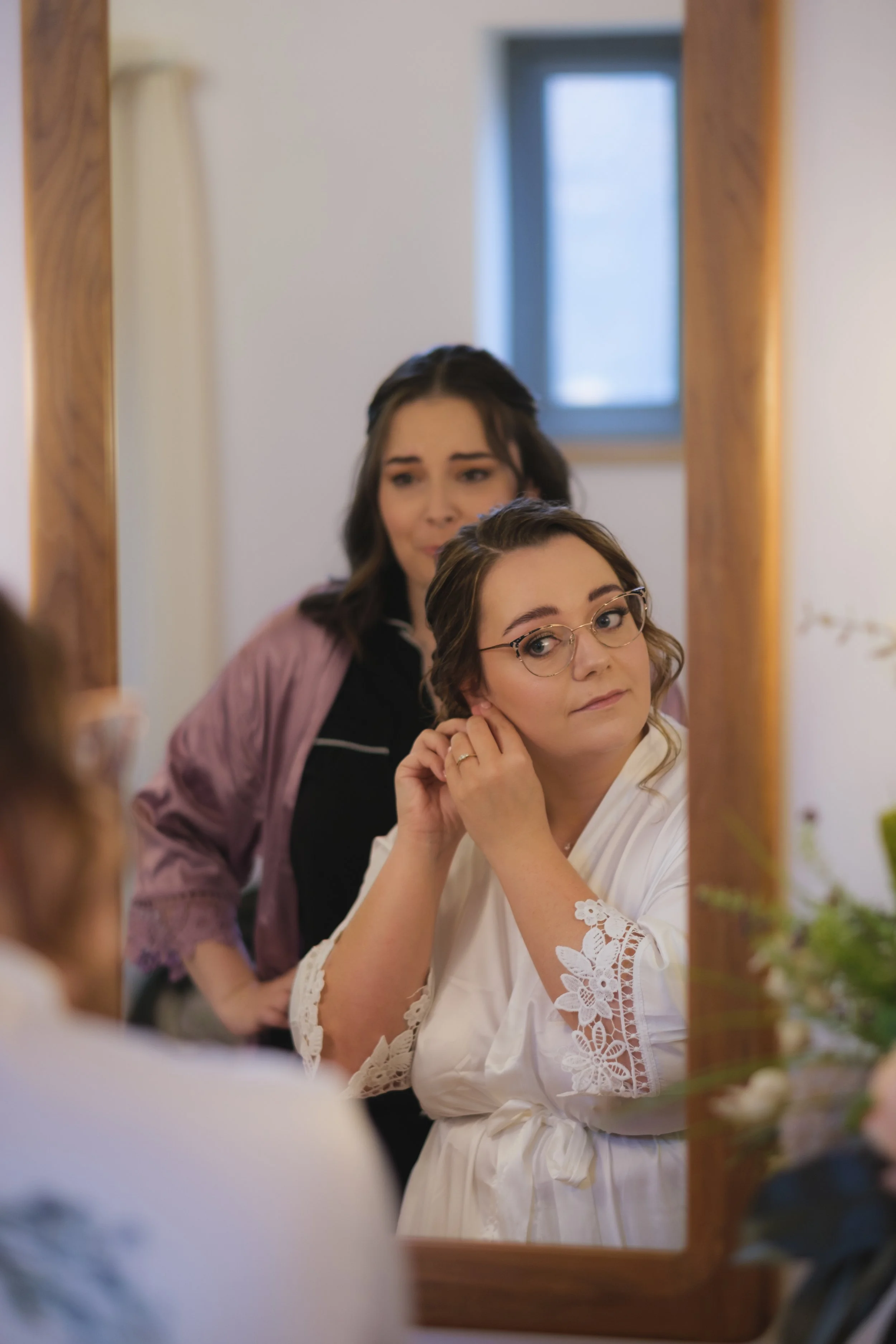 Bride in a white robe wearing earrings in front of a mirror, with a bridesmaid in a pink robe standing behind her, indoors.