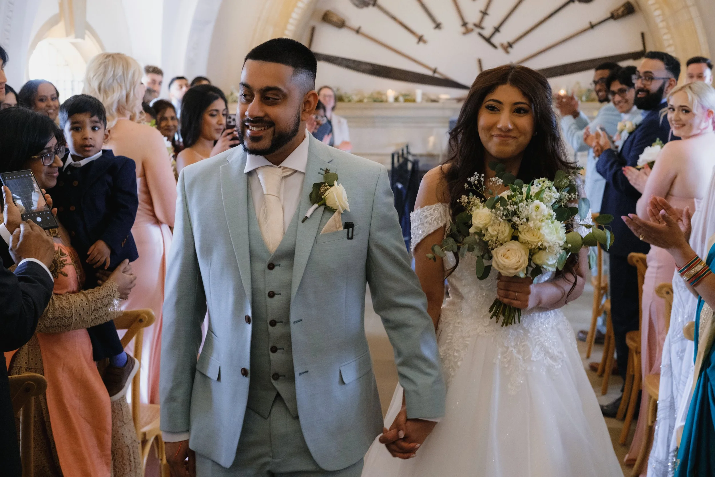 A bride and groom holding hands walking down the aisle at their wedding ceremony, surrounded by guests who are clapping and taking photos. The bride is wearing a white gown and holding a bouquet of white flowers, while the groom is in a light blue su