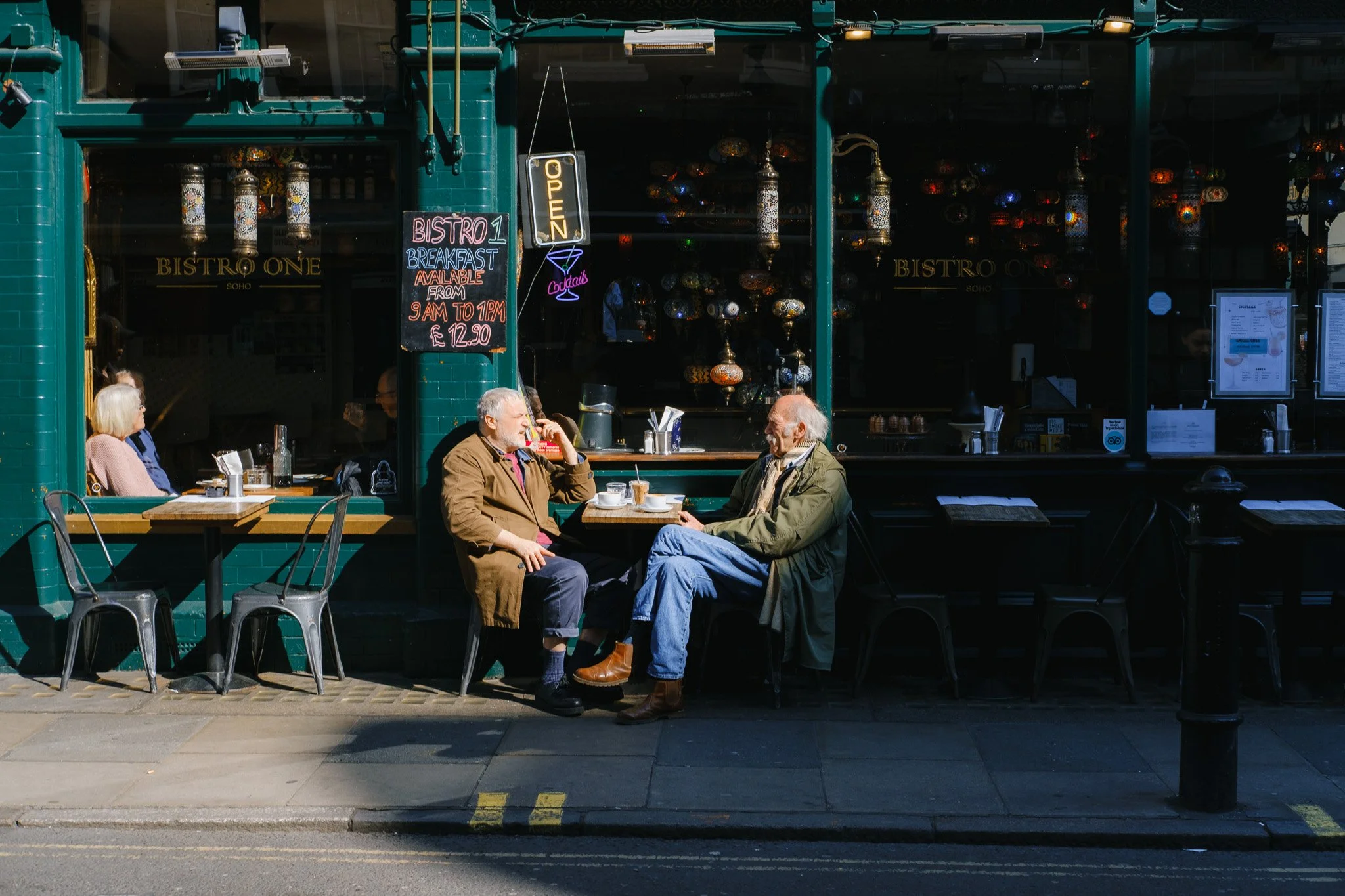 Two men sitting at a small outdoor table outside a bistro, engaged in conversation, with coffee cups on the table. A woman sits alone at another table by the window of the bistro. Signs and decorations are visible in the background.