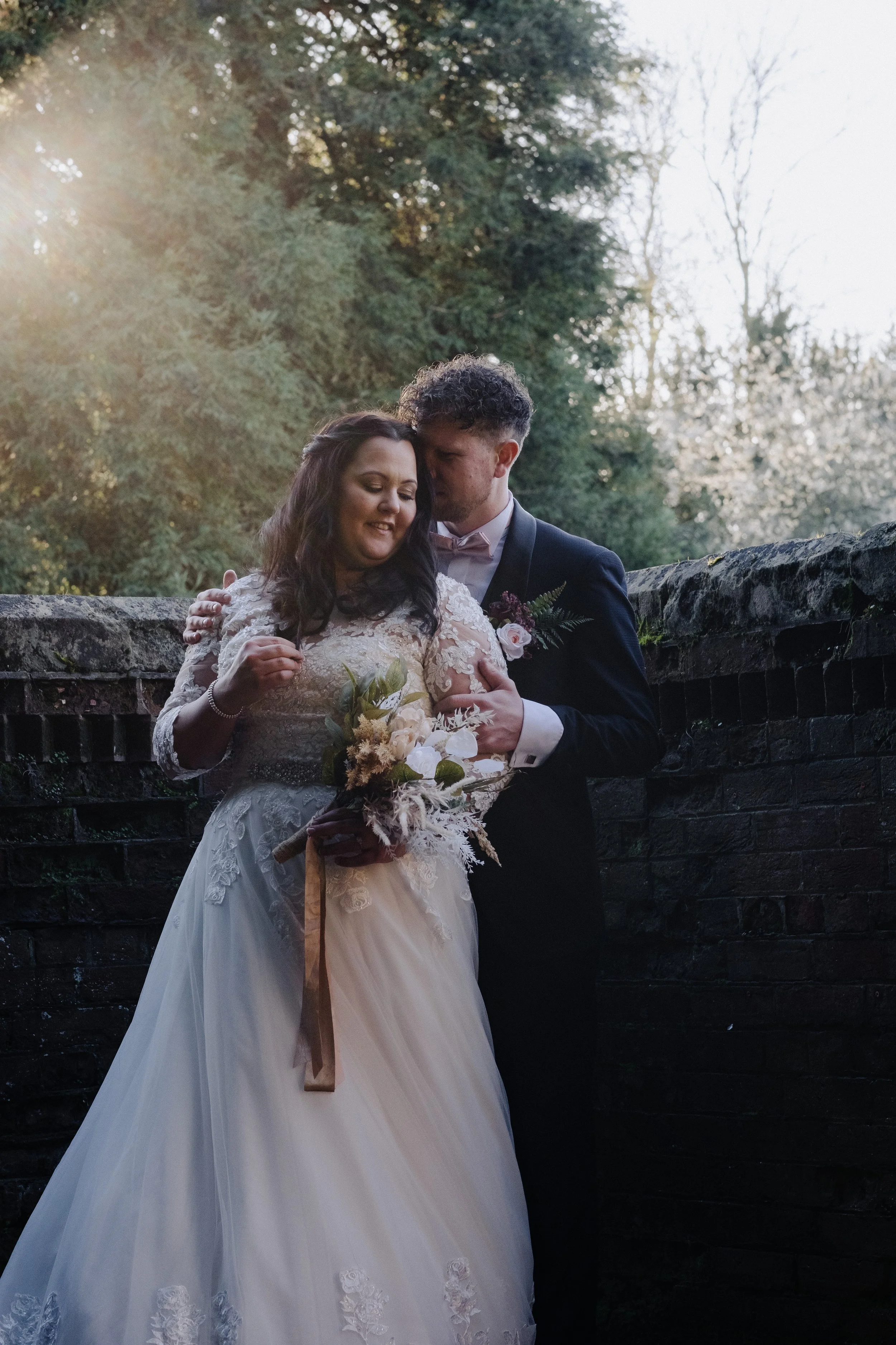 Bride and groom embracing outdoors, bride in a white gown holding a bouquet, groom in a suit.