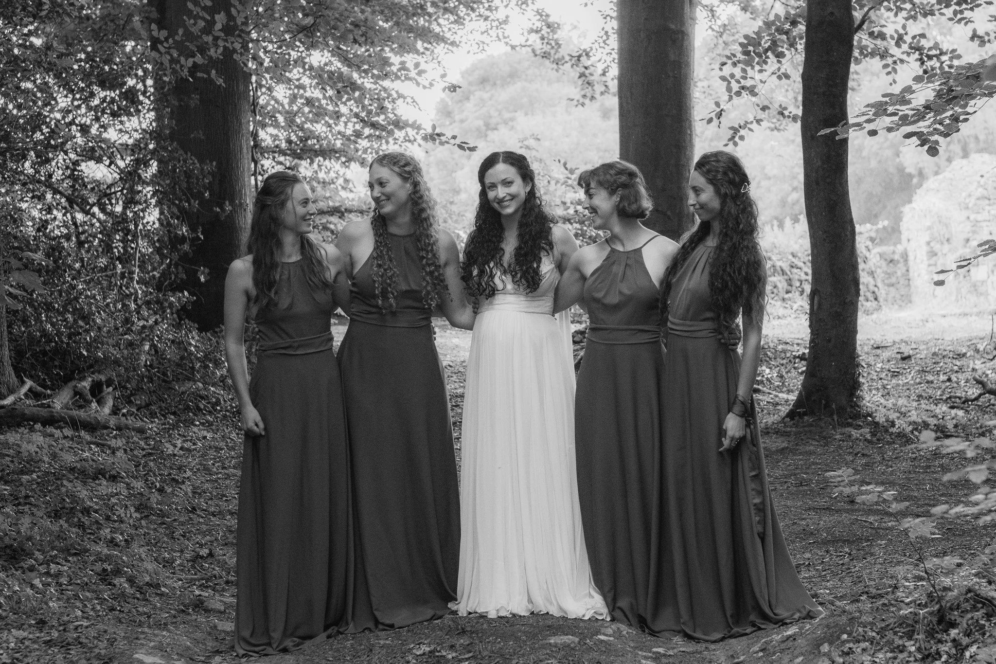 A group of five women in dress standing in a wooded outdoor setting, smiling and holding each other.