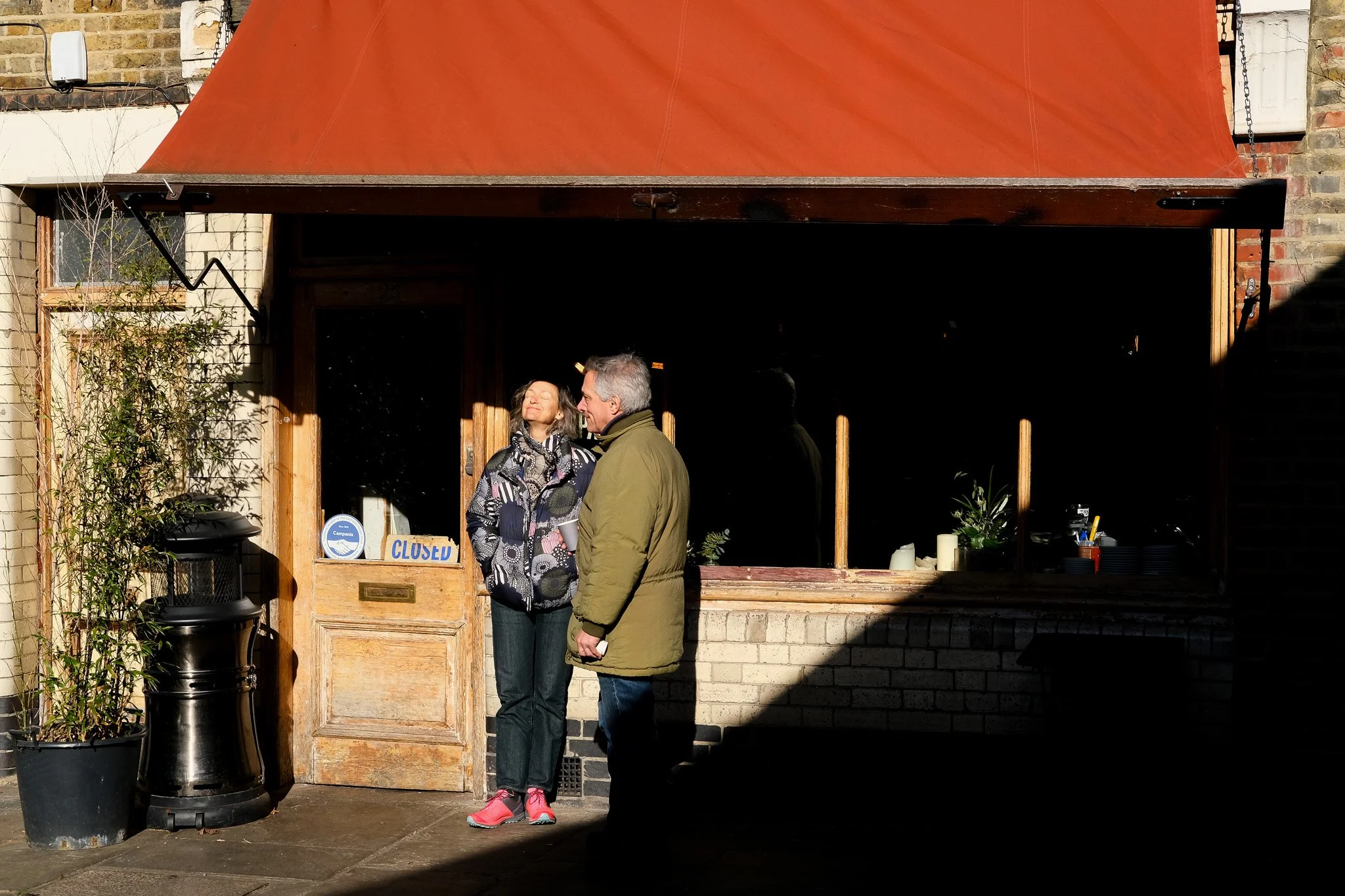 Two people standing outside a closed café under a red awning, one person facing upward towards the sun, the other looking at them, against a brick wall backdrop.