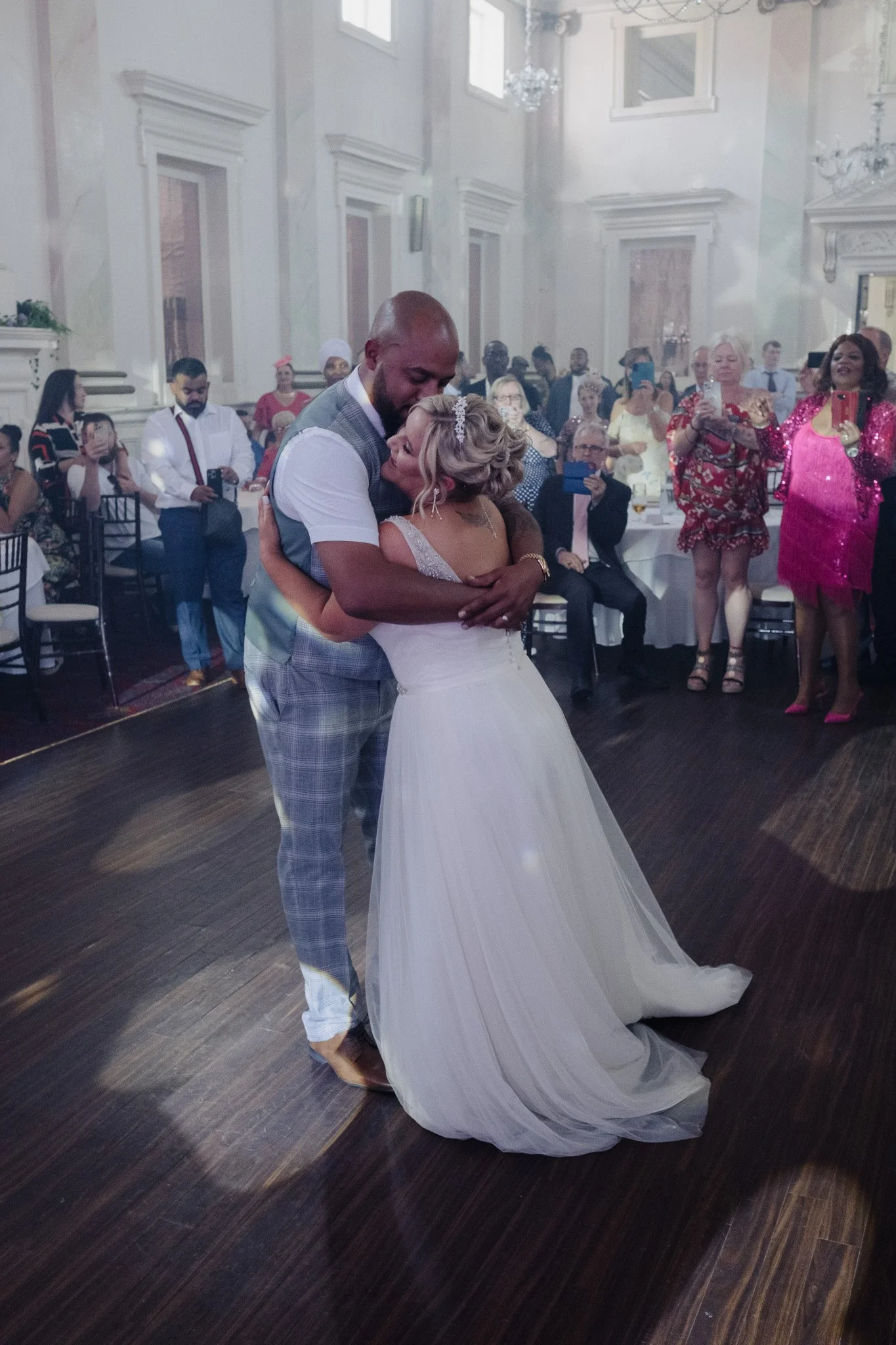 A bride and groom embracing on a dance floor during a wedding, surrounded by guests watching and taking photos. The bride is wearing a white gown and the groom is in a suit. The venue is elegantly decorated with chandeliers and large windows.