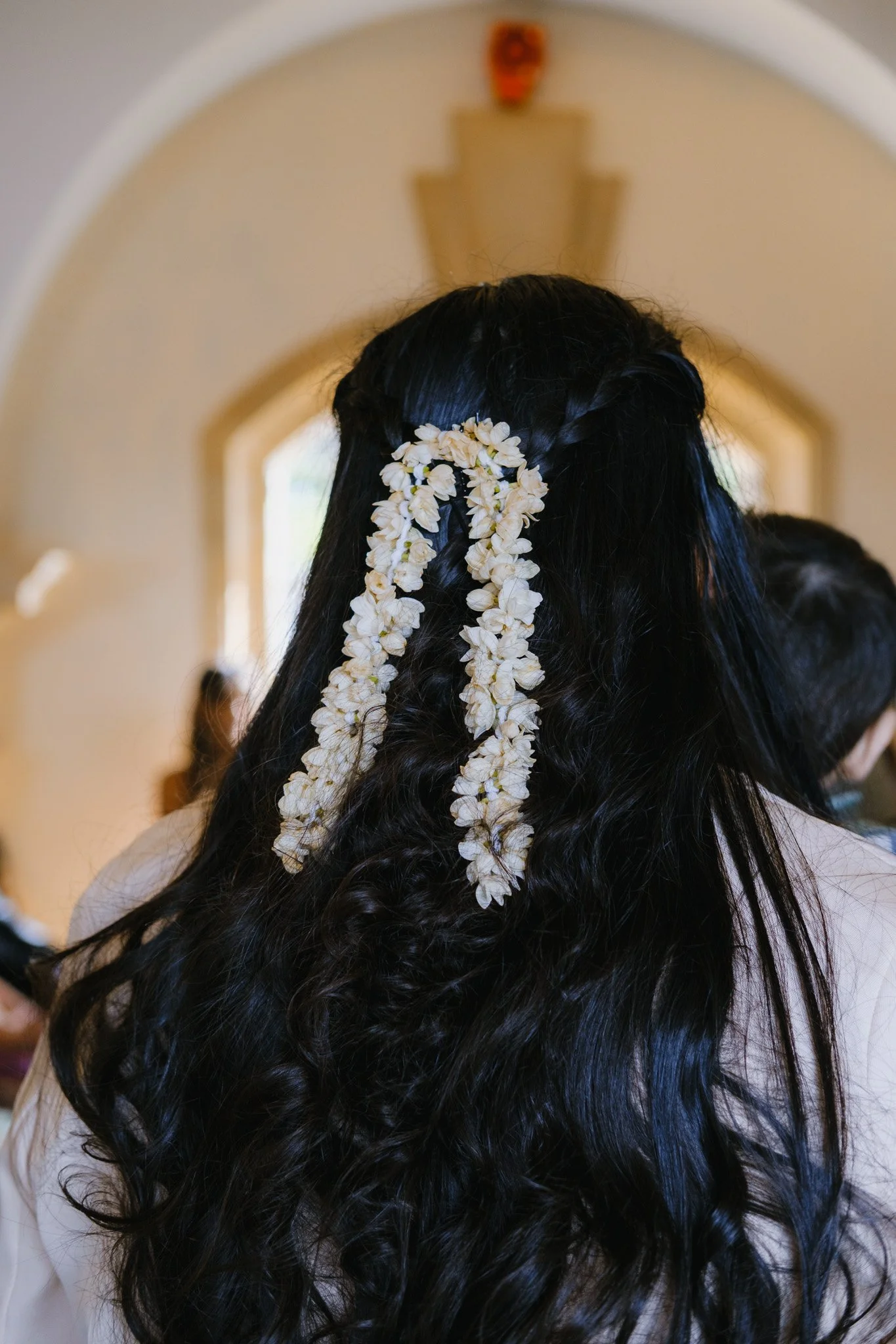 Woman with long dark hair adorned with white flower garlands, back view, indoors.