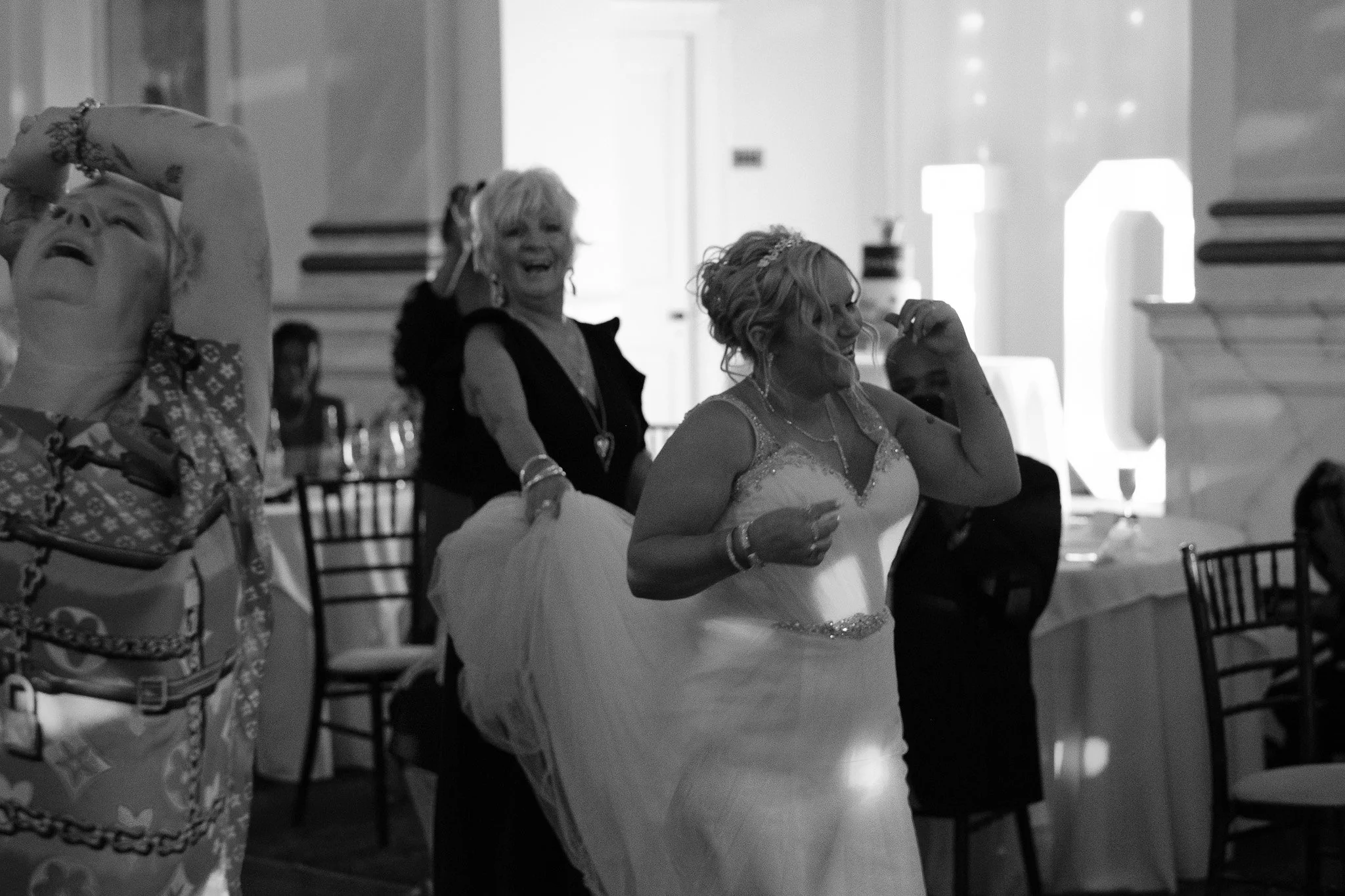 Black and white photo of three women dancing at a wedding reception in a banquet hall, with one wearing a wedding dress.