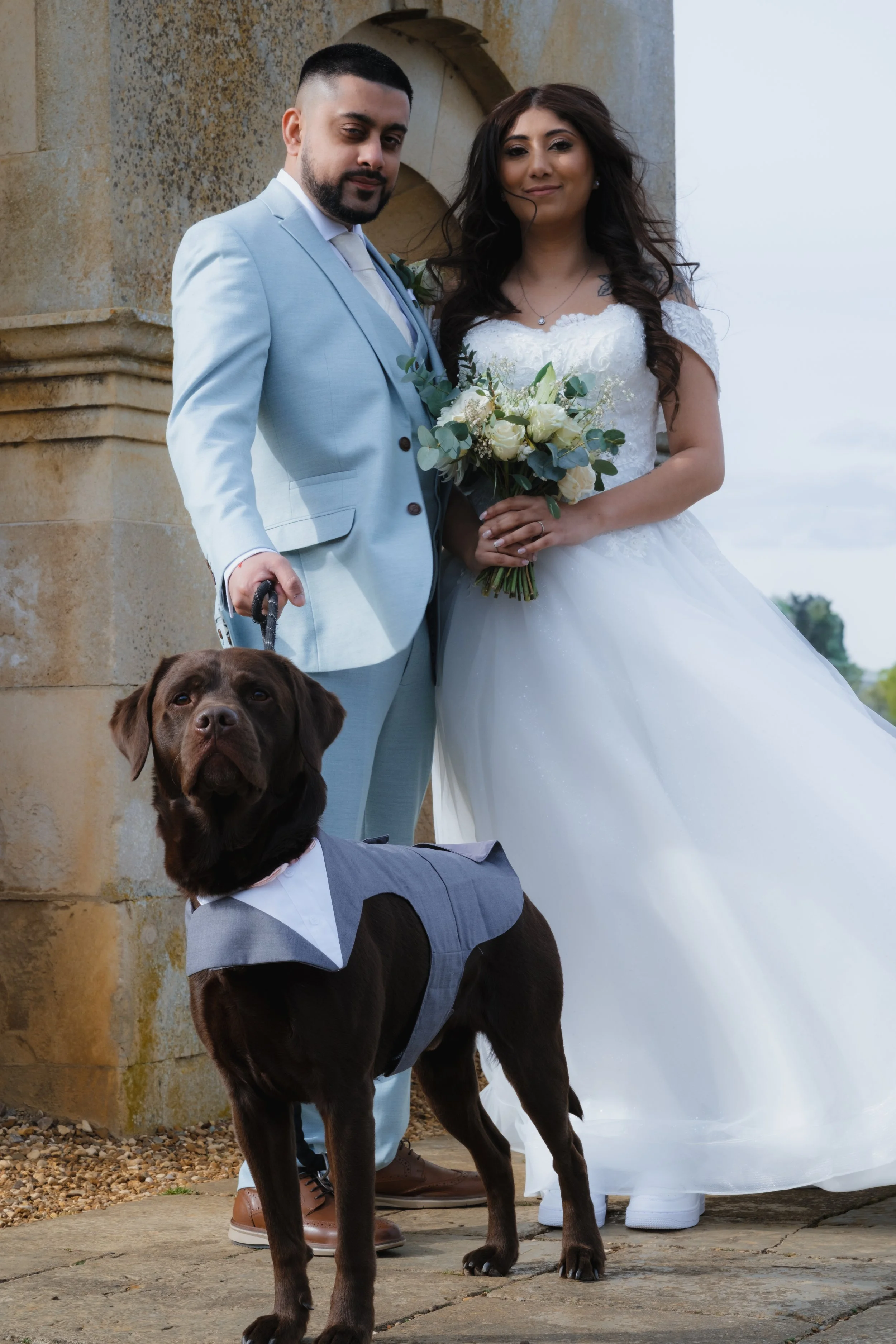 A bride and groom standing with a chocolate Labrador dressed in a formal outfit. The bride is holding a bouquet.