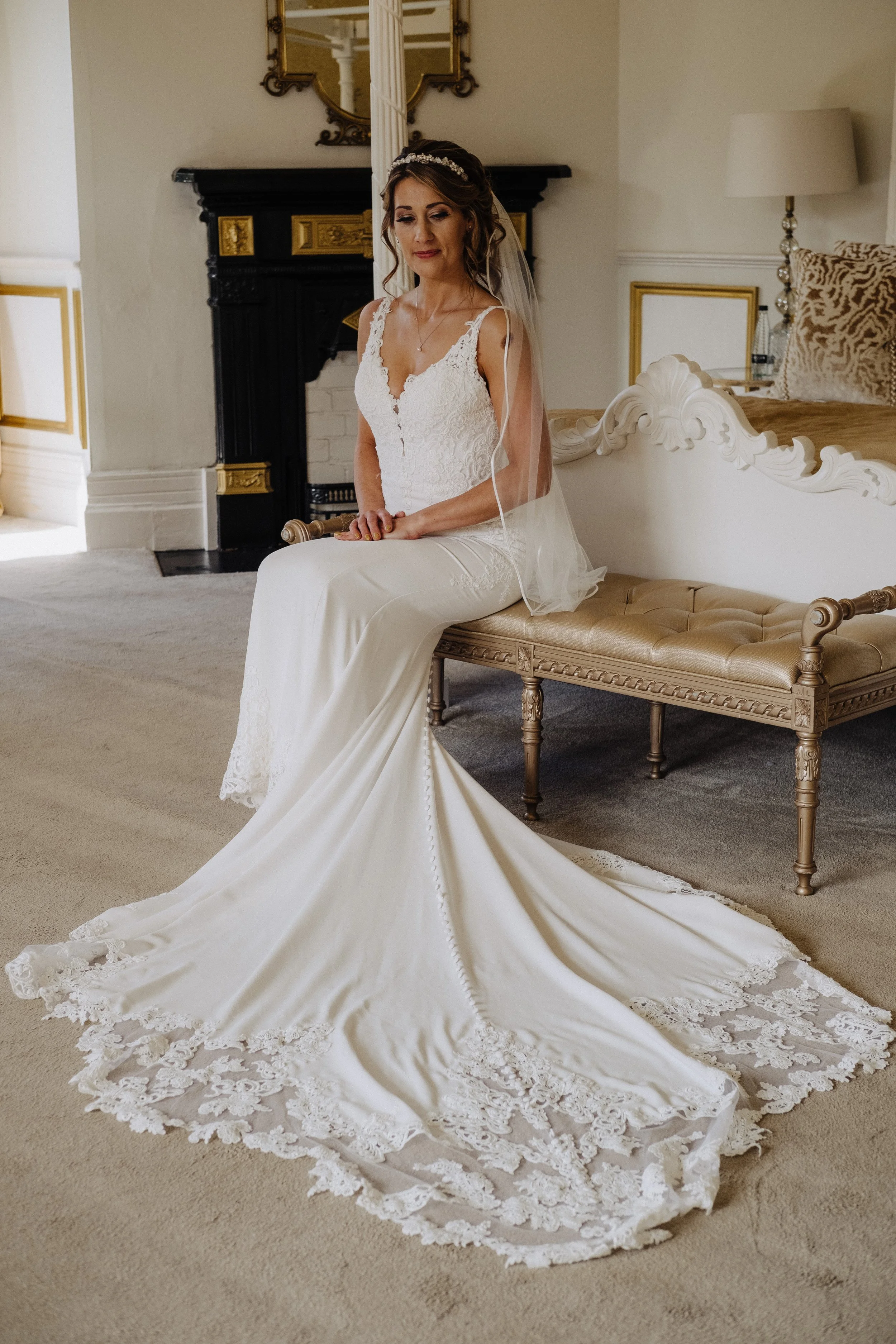 A bride sitting on an elegant bench, wearing a white lace wedding dress with a long train and veil, in a vintage-style room with a fireplace and ornate mirror.