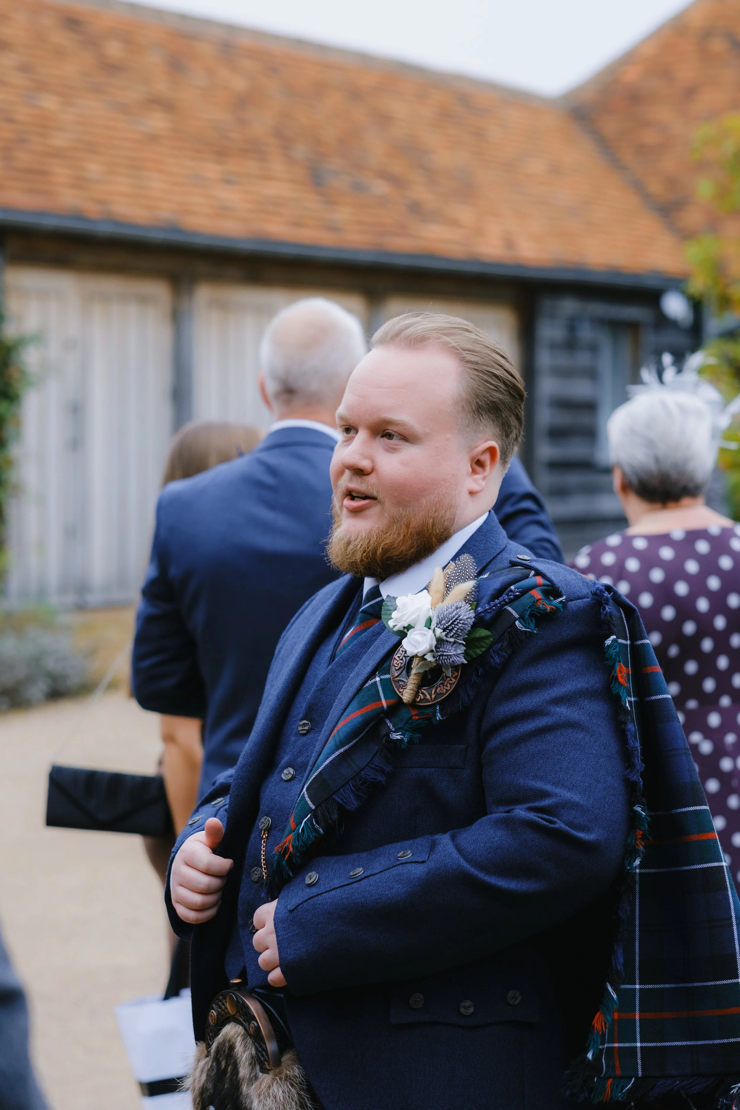 Man in a blue formal outfit with a plaid sash at an outdoor event with people in the background.