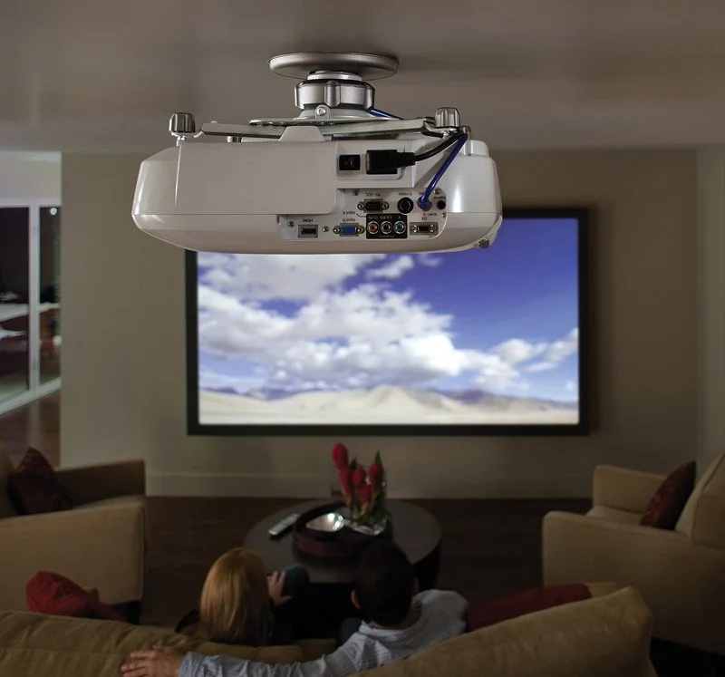 A ceiling-mounted projector displaying a landscape image of a desert with mountains and a blue sky with clouds, inside a living room with two children sitting on a sofa watching the screen.