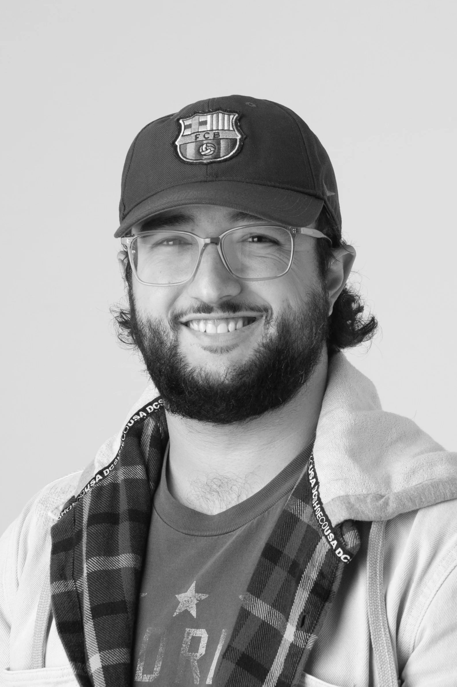 Black and white photo of a man with glasses and a beard, wearing a baseball cap and a T-shirt from The North Face, smiling at the camera.