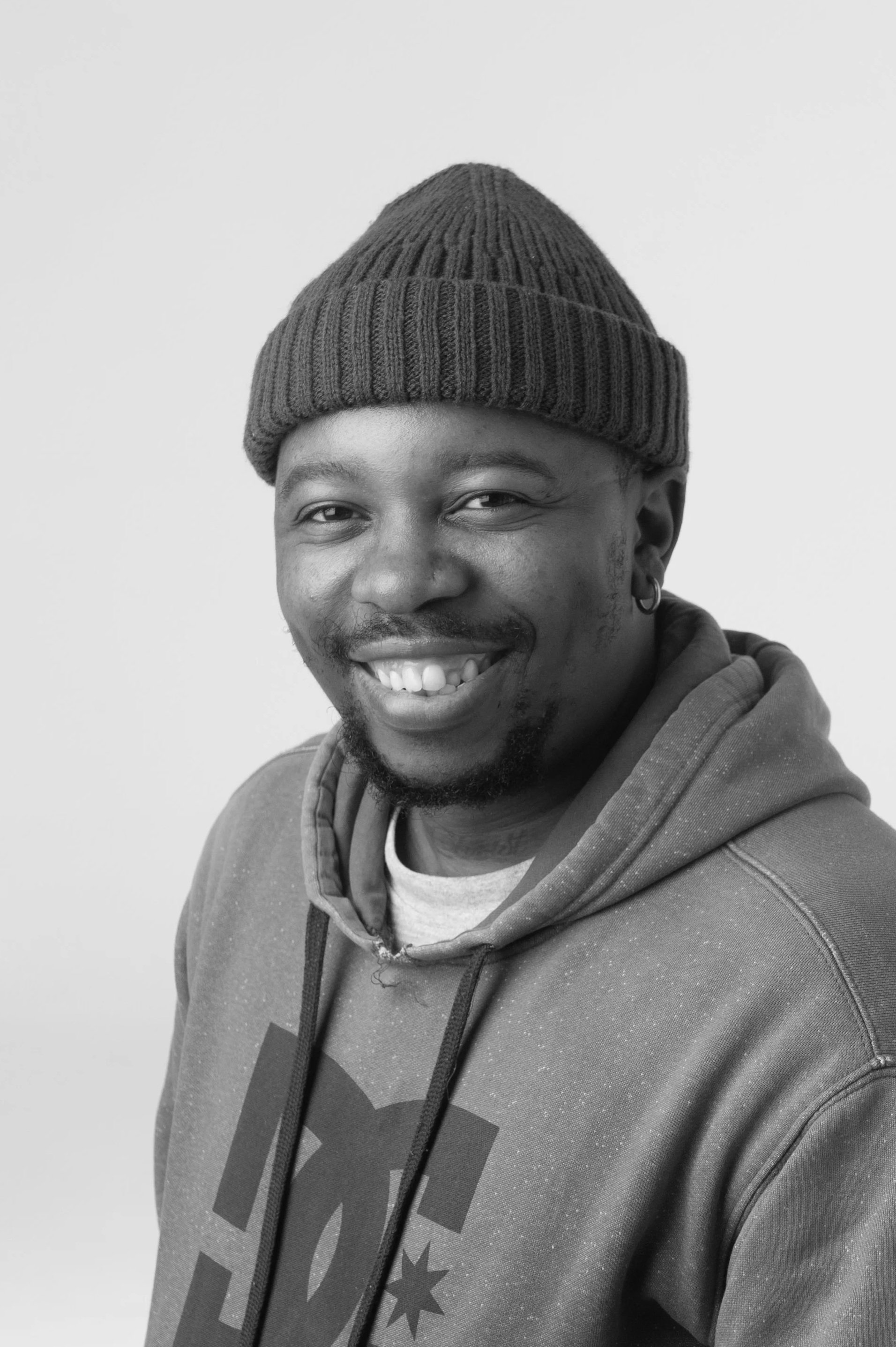 Black and white portrait of a smiling man wearing glasses, a cap, hoop earrings, and a light-colored t-shirt.