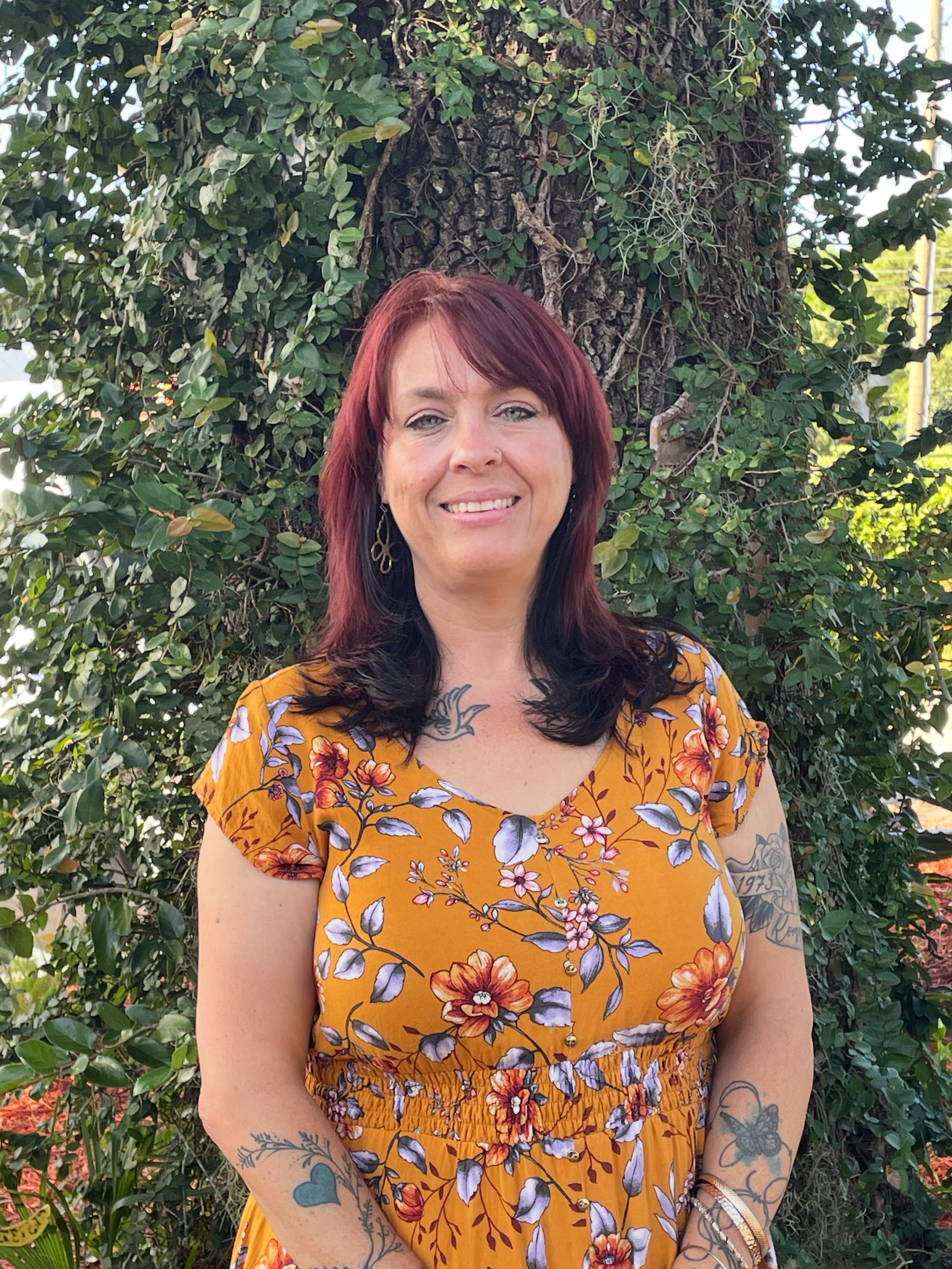 Ms. Salli, A woman with long red hair smiling, wearing a black floral top with purple, yellow, and white flowers, gold jewelry, and a nose ring, standing against a light blue background